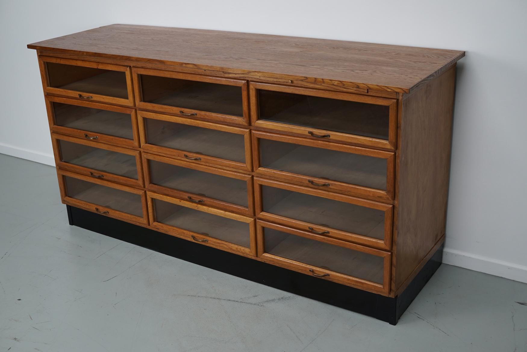 This haberdashery cabinet was produced during the 1950s in the Netherlands. This piece features 12 large drawers in oak with glass fronts and brass handles. It was originally used in a luxury warehouse in the city of Amsterdam. The interior