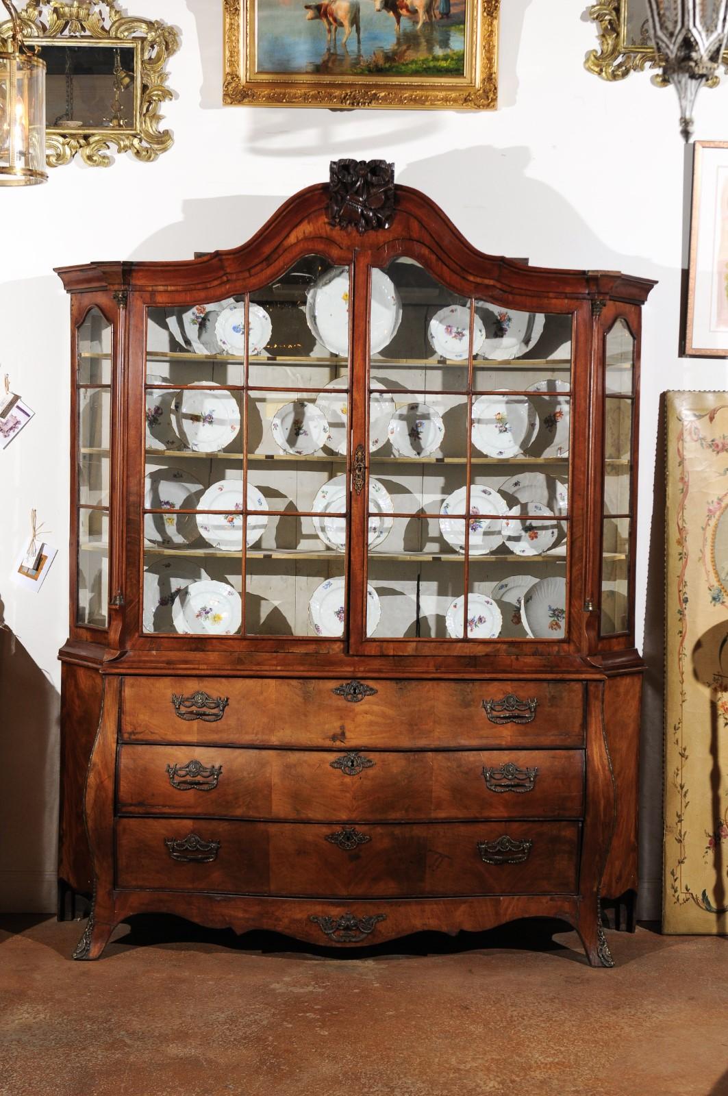 A Dutch Rococo period walnut vitrine cabinet from the late 18th century, with hand carved hunting trophy, glass doors and bombé chest. Born in the Netherlands in the later years of the 18th century, this exquisite Rococo cabinet features a