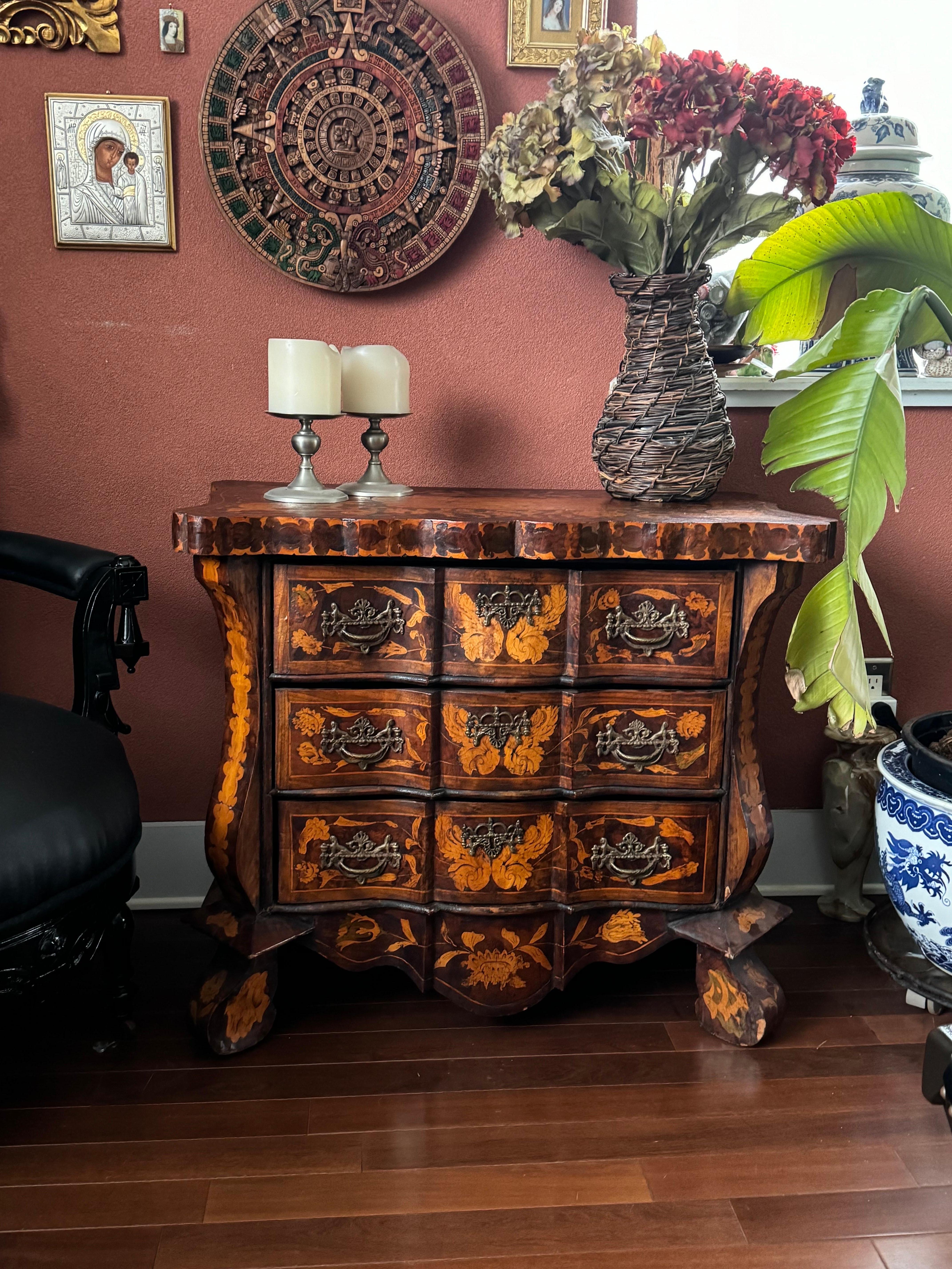 This is a Dutch Rococo style commode, featuring intricate satinwood marquetry on a burl mahogany background. The commode has a serpentine front, ornate with floral and foliage inlays, and is fitted with metal handles on the drawers. The curvature
