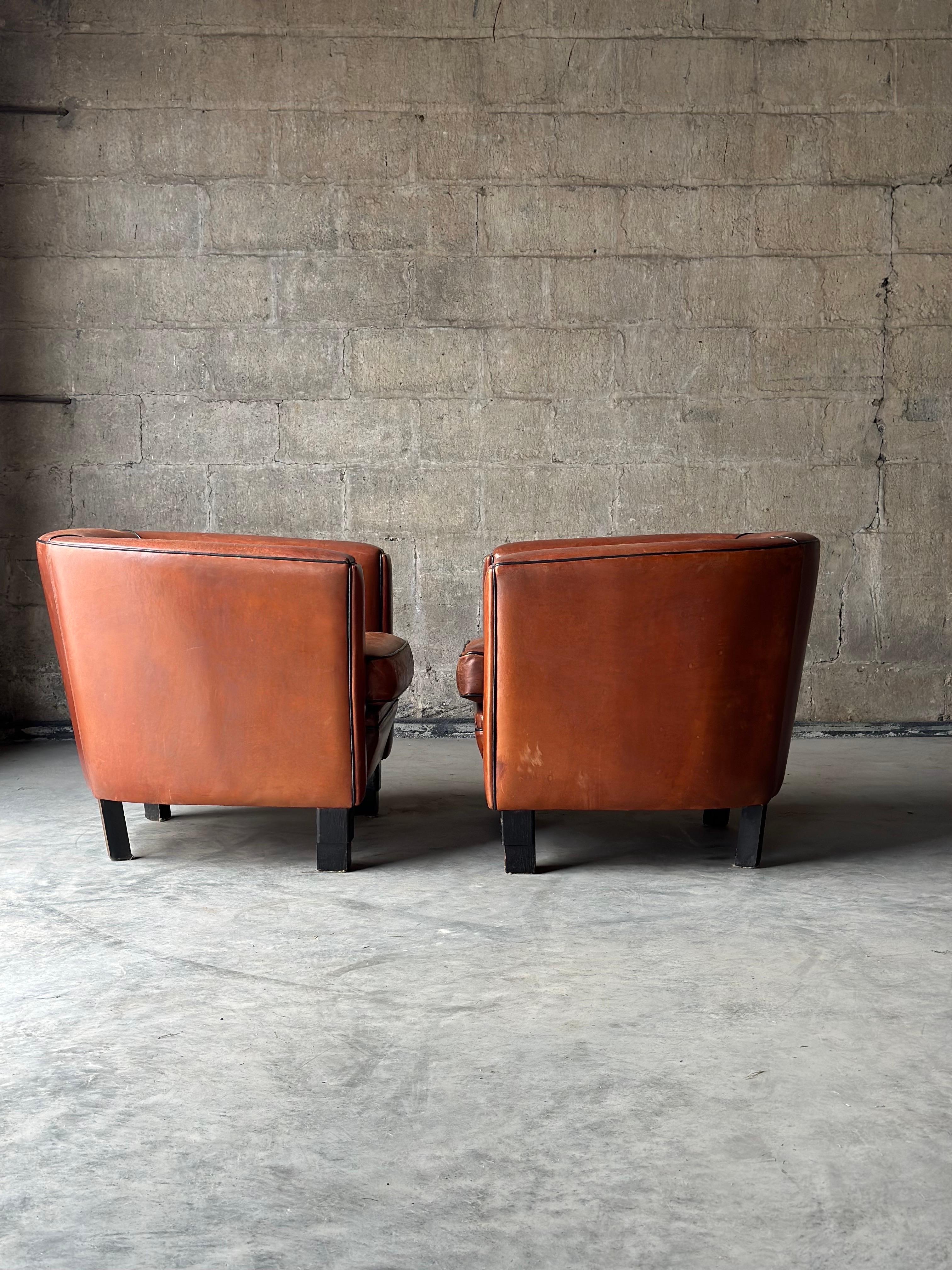 Perfect patina on this cool pair of Dutch club chairs. Cognac colored sheep’s leather is framed with black piping that matches black wooden feet.