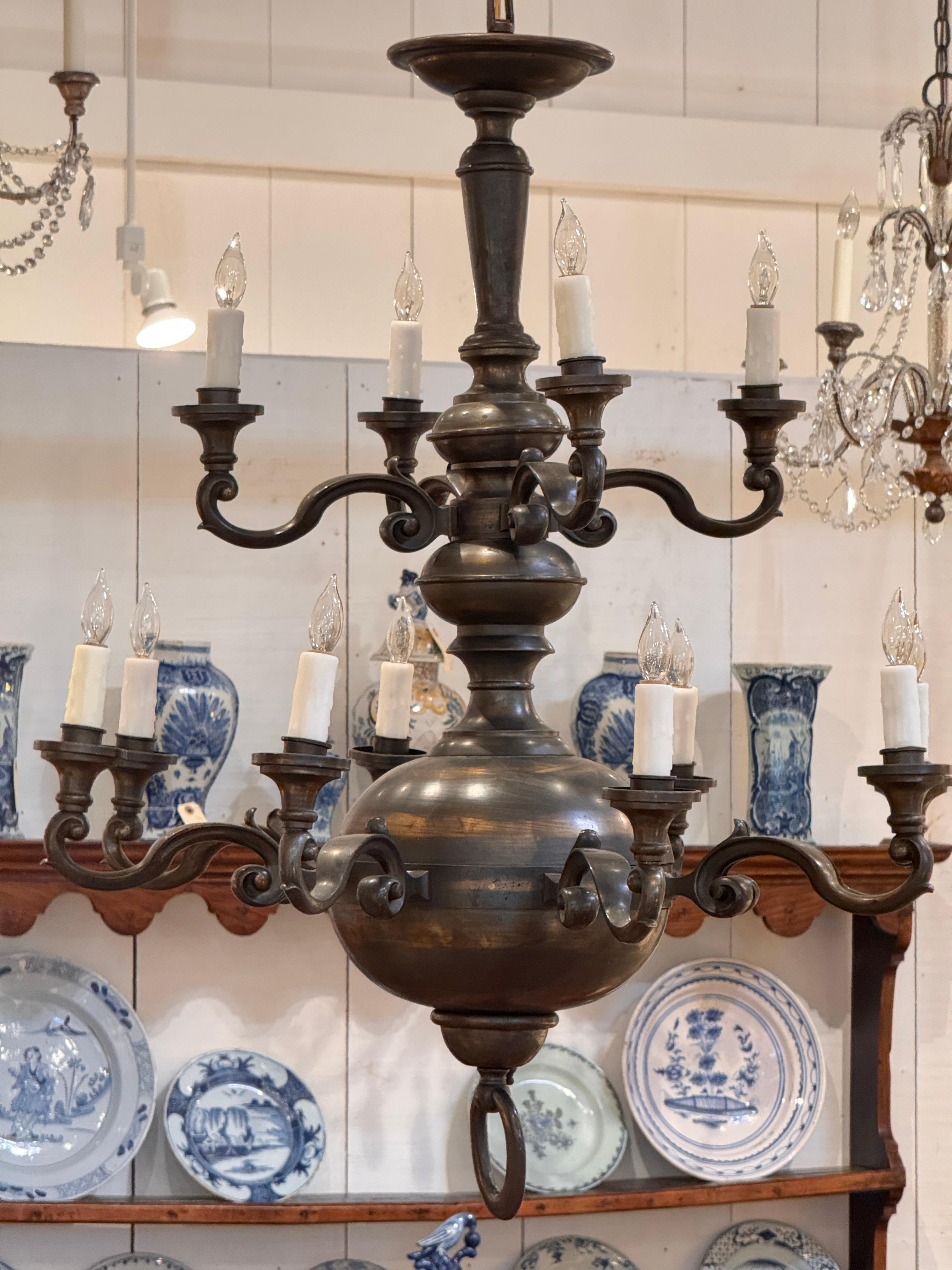 Lampadario a dodici luci in stile olandese, con finitura in ottone patinato, bracci graziosamente arcuati e forma a doppio binario. La sfera centrale presenta un'usura sottile e un carattere coerente con l'età.