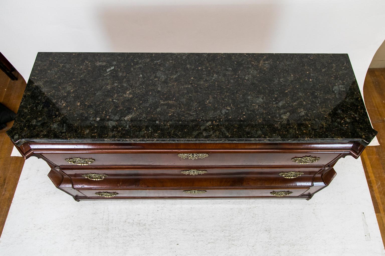 This three-drawer granite top chest has a dark stone top, which is later. There are elaborate handles which were individually hand cast but are not original. The top frieze is concave. The middle drawer has a concave shape above a straight drawer.