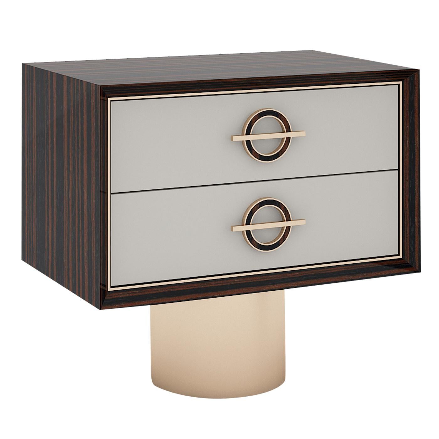 Distinguished by a geometric structure, this polished nightstand showcases a brass-finished cylinder sustaining its wooden top that includes two grey-lacquered drawers. Two circular handles with a horizontal brass-finished metal segment complete the