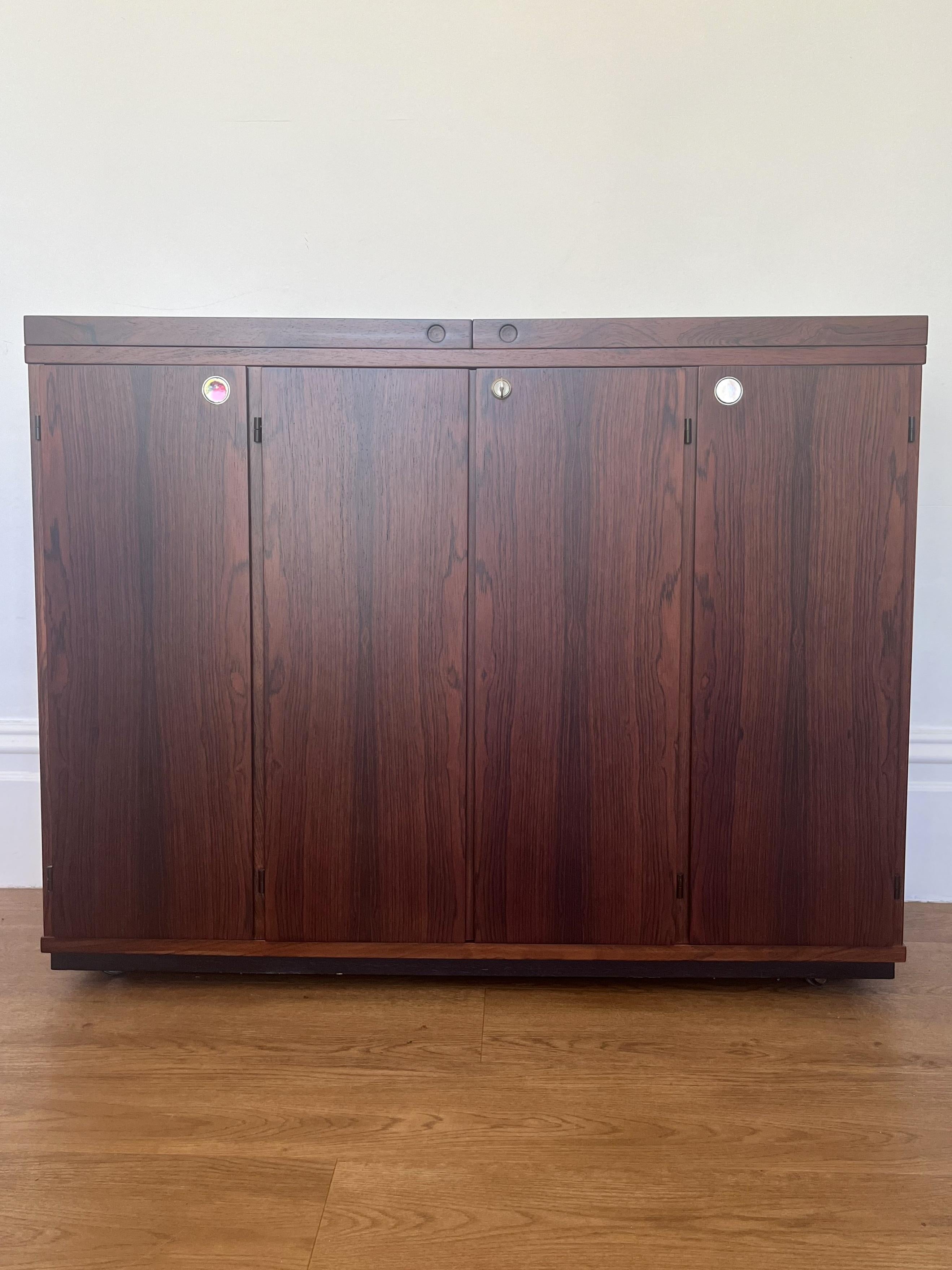 Dyrlund Rosewood drinks cabinet on castors.

The cabinet has an extending pull out top with black formica bar area.  2 single and 1 lockable central double door.  The interior has ample storage space which includes a total of 4 drawers and slide out