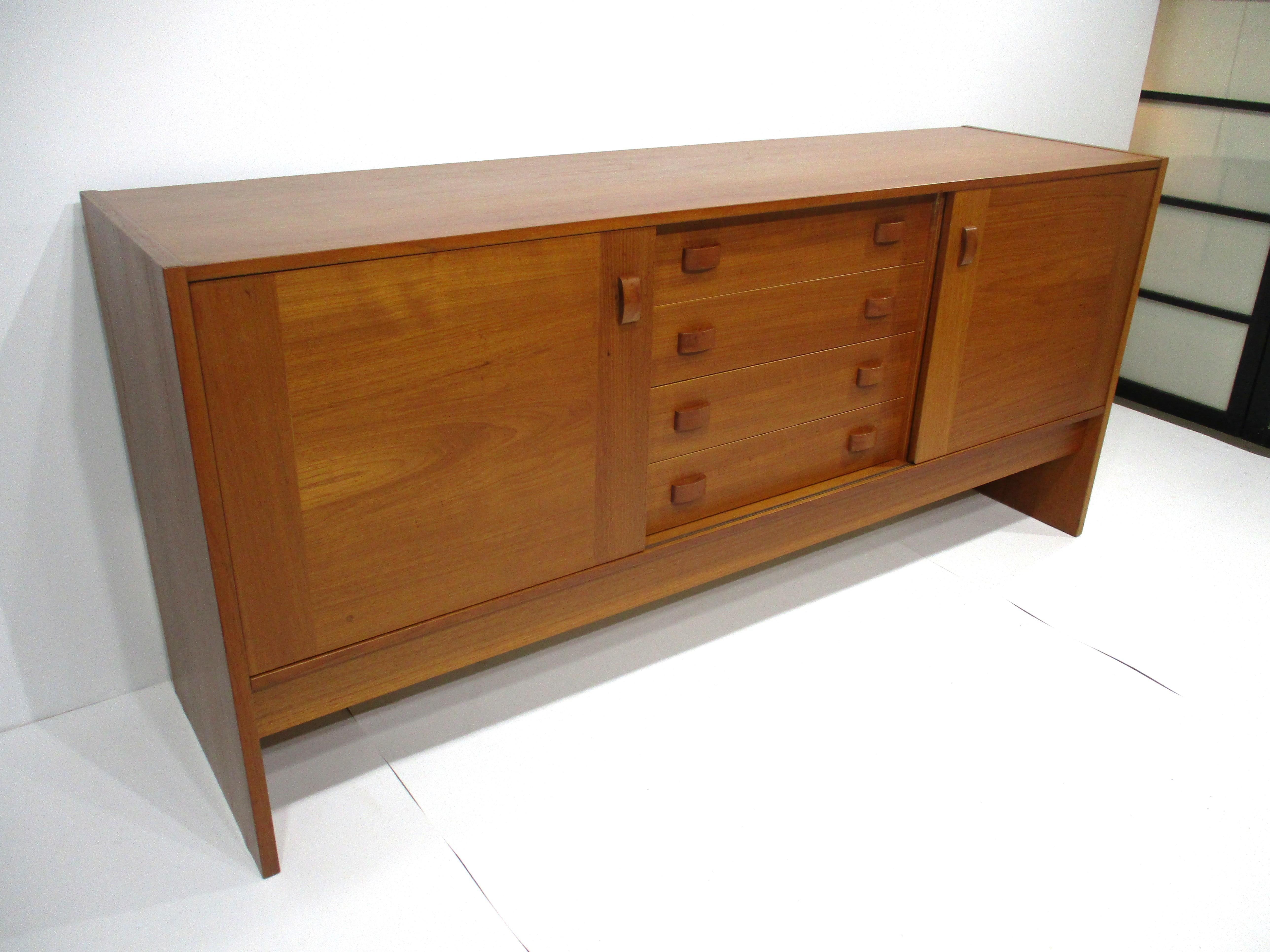 Una grande credenza in legno di teak con quattro sottili cassetti centrali e, su ciascun lato, un'anta scorrevole con due ripiani regolabili. Le maniglie in teak abbinate consentono un'apertura e una chiusura agevoli e un ampio spazio di