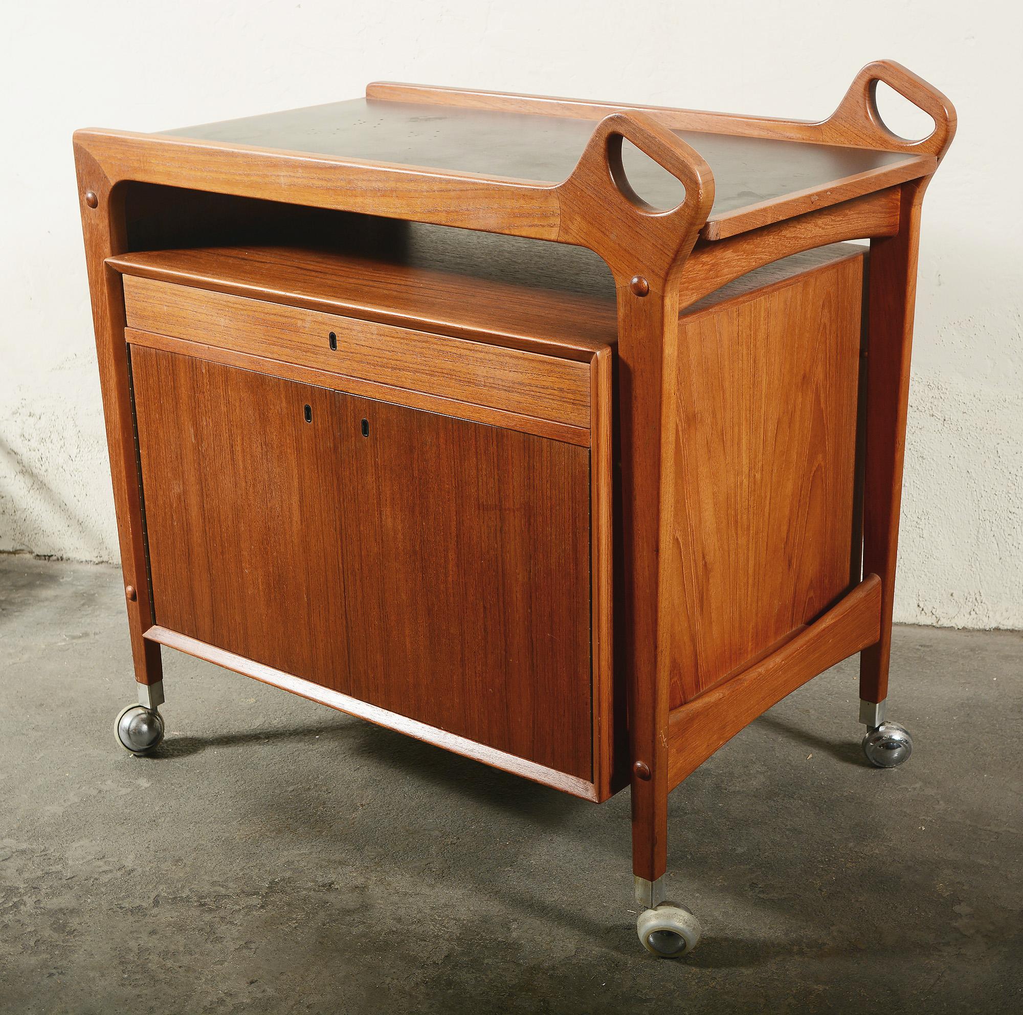 Teak bar cart designed by Johannes Andersen for Dyrlund. This cart has a drop leaf top that when raised makes the top 57 1/8 inches wide. An enclosed cabinet floats below the top. It consists of a divided drawer and two cabinet doors. One side of