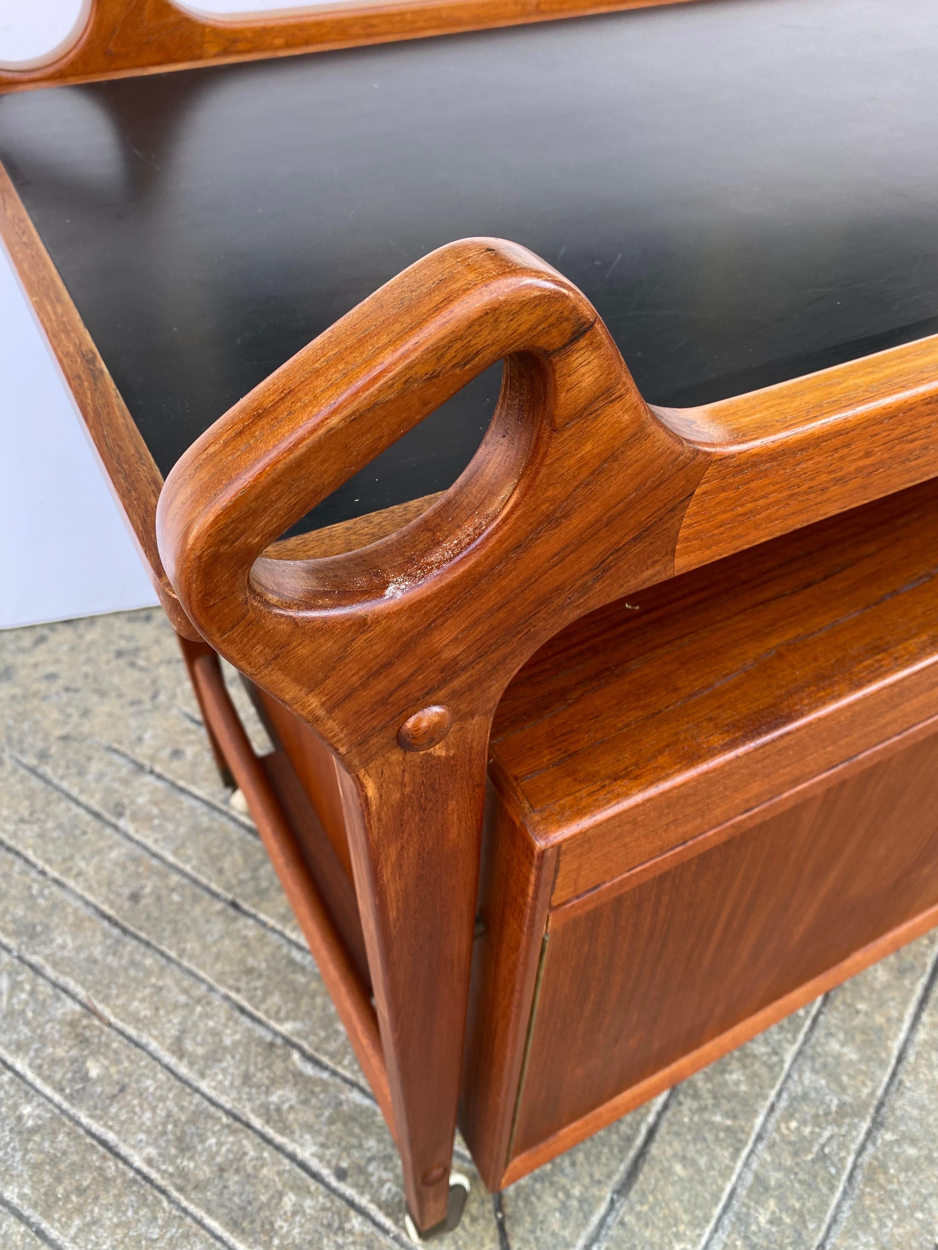 Dyrlund Teak Bar with locking doors and drawer. Top lifts up and slides over to add additional work surface. Top surface is black formica. 2 carved handle holds make it easy to move around a room. Casters move easily. Label on underside.