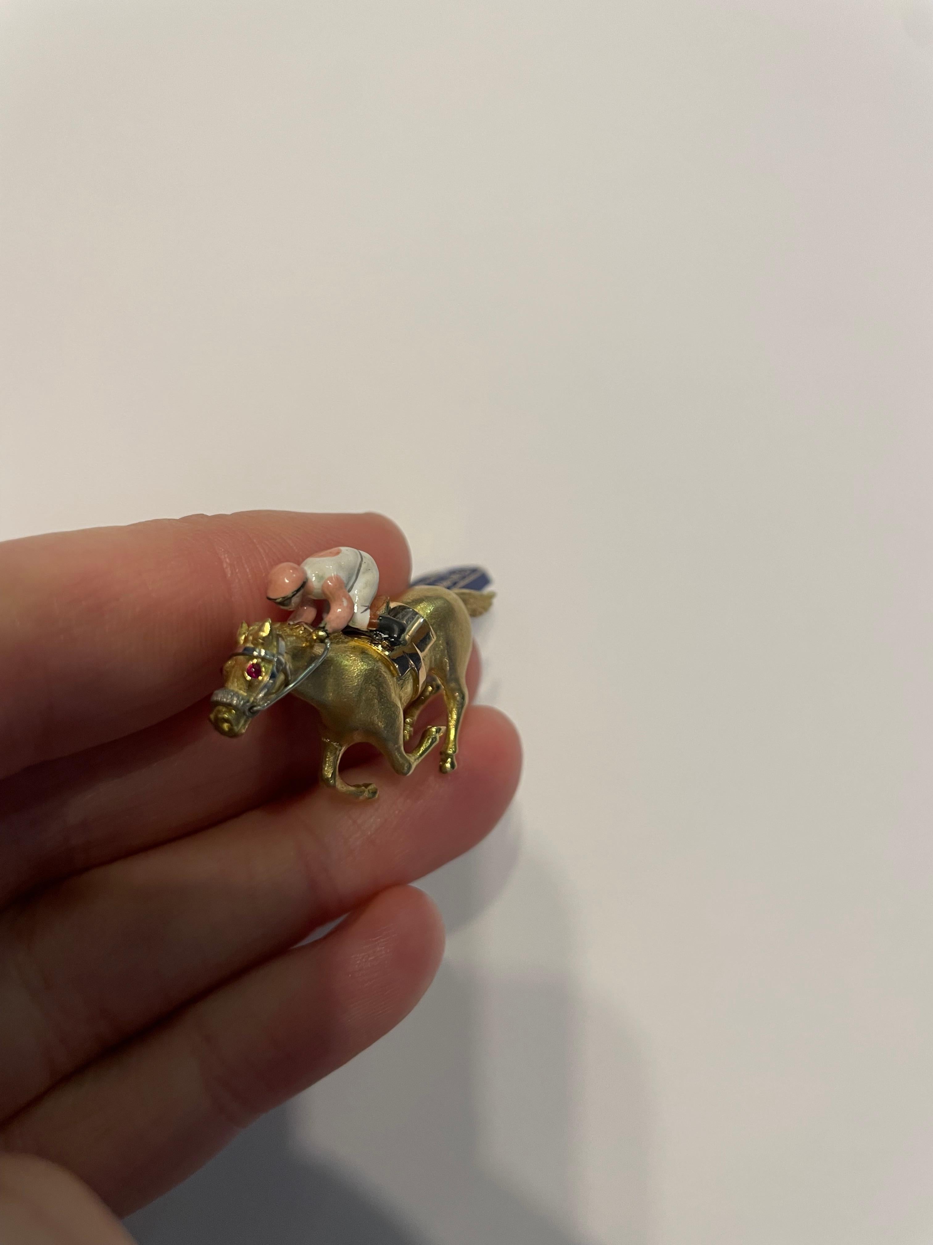 Spilla per cavalli da corsa realizzata a mano da E. Wolfe & Company in oro giallo 18 carati. Il cavallo è incastonato con occhi di rubino cabochon e il cavaliere è smaltato di rosa e bianco. La spilla pesa 14.30 grammi. Il retro è dotato di una