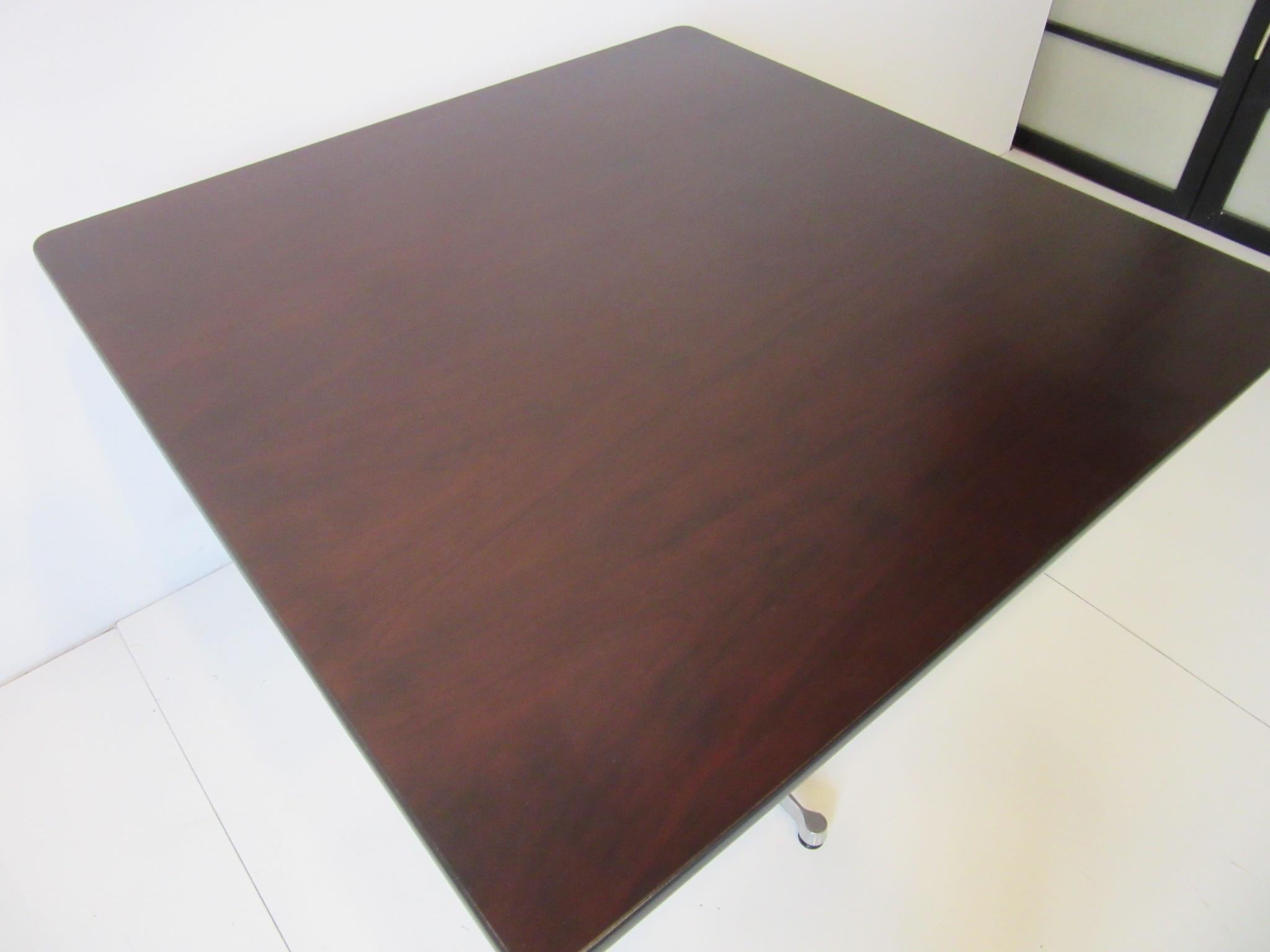 A darker rosewood toned square Eames aluminum group smaller sized dining table with rolled black rubber edge, satin black metal post mounted to a cast aluminum star base with adjustable feet. Retains the original manufactures label from the Herman