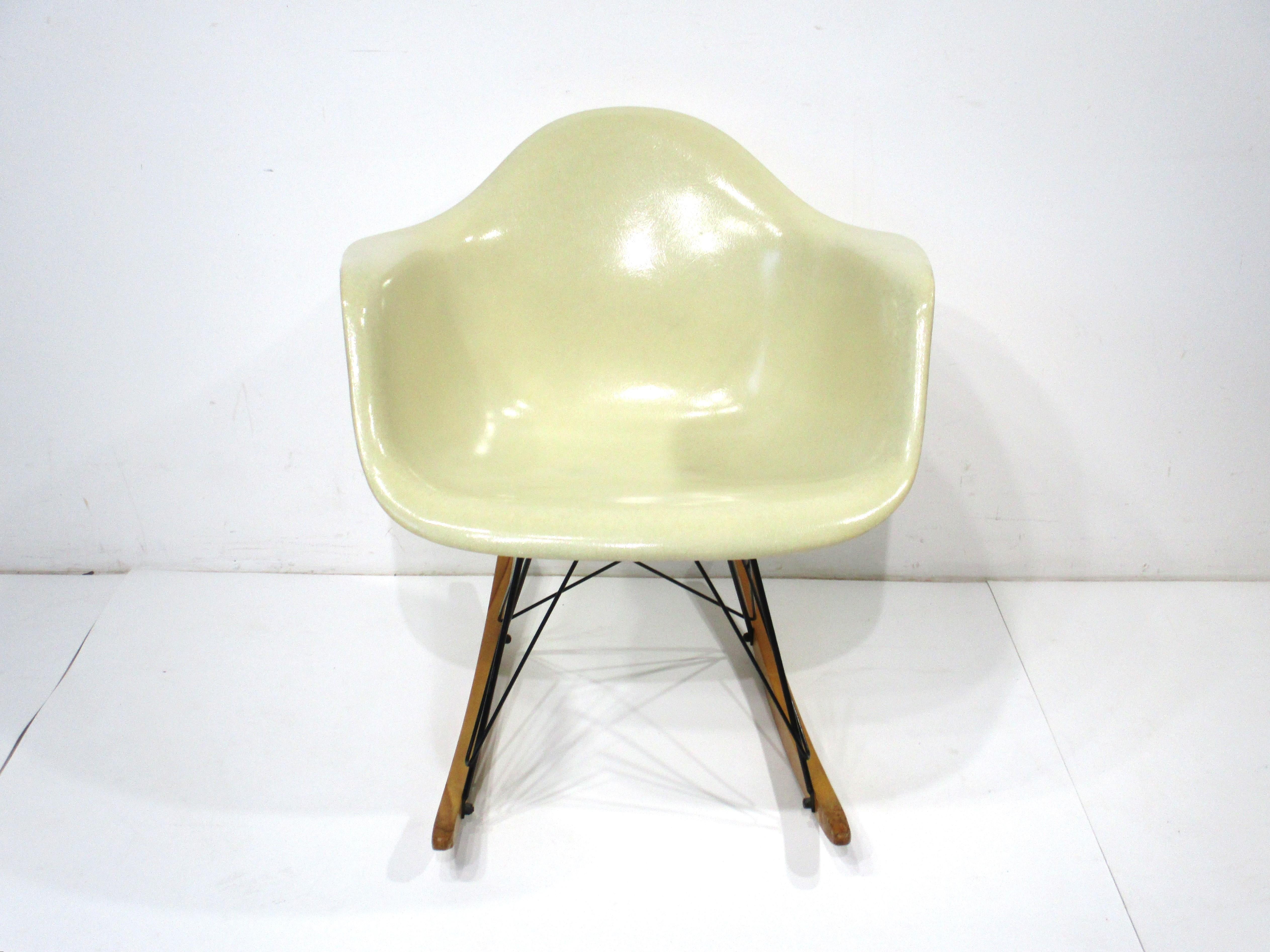 A very nice iconic Mid Century designed molded fiberglass rocking arm shell chair in a parchment color . The satin black base is constructed in metal rods with birch wood runners giving the piece great contrast . Designed by Ray and Charles Eames