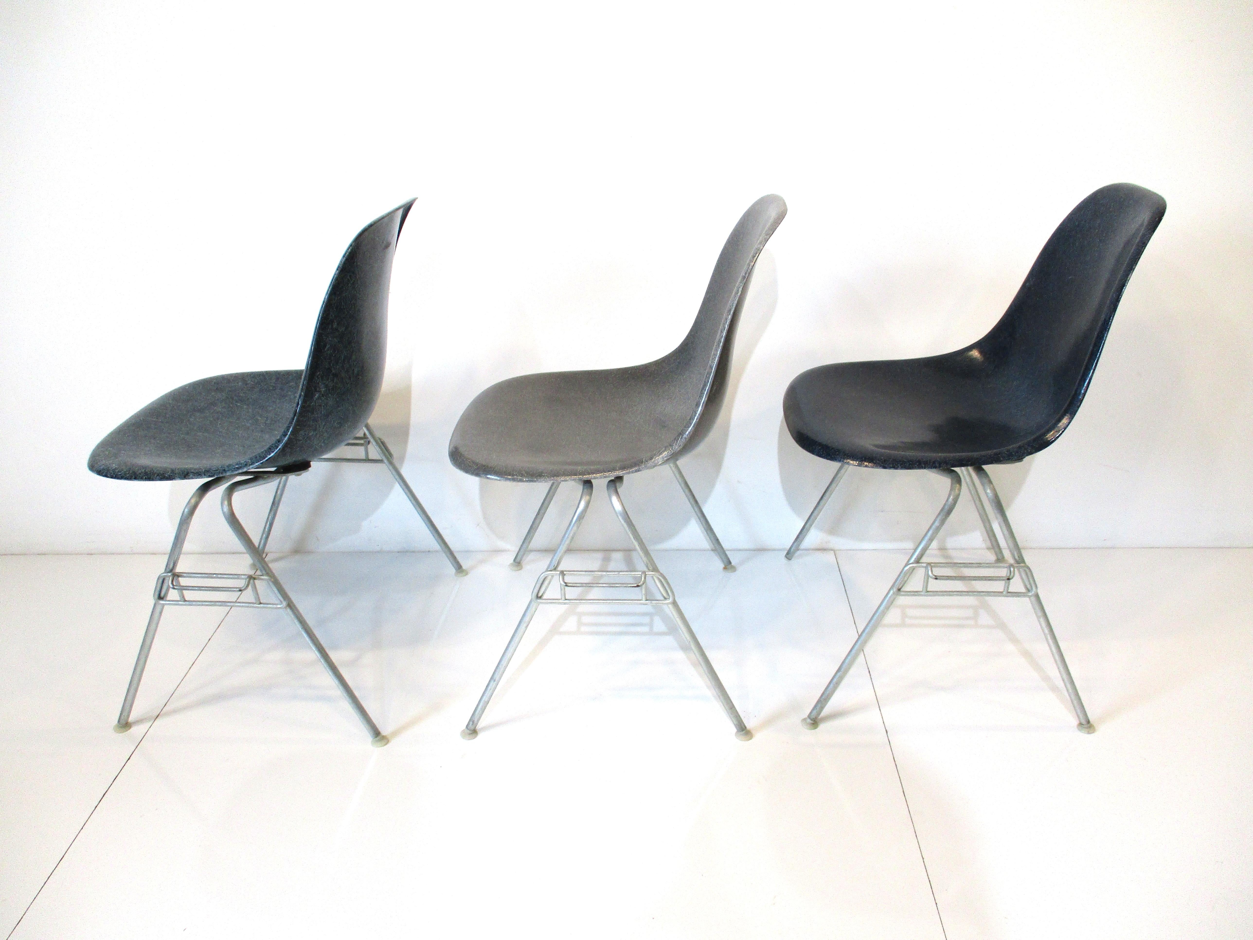 A set of three fiberglass DSS stacking dining chairs with zinc H bases and nylon foot pads . The colors are gray , medium blue and dark blue with the Herman Miller molded manufactures mark and ink stamp to the bottoms.