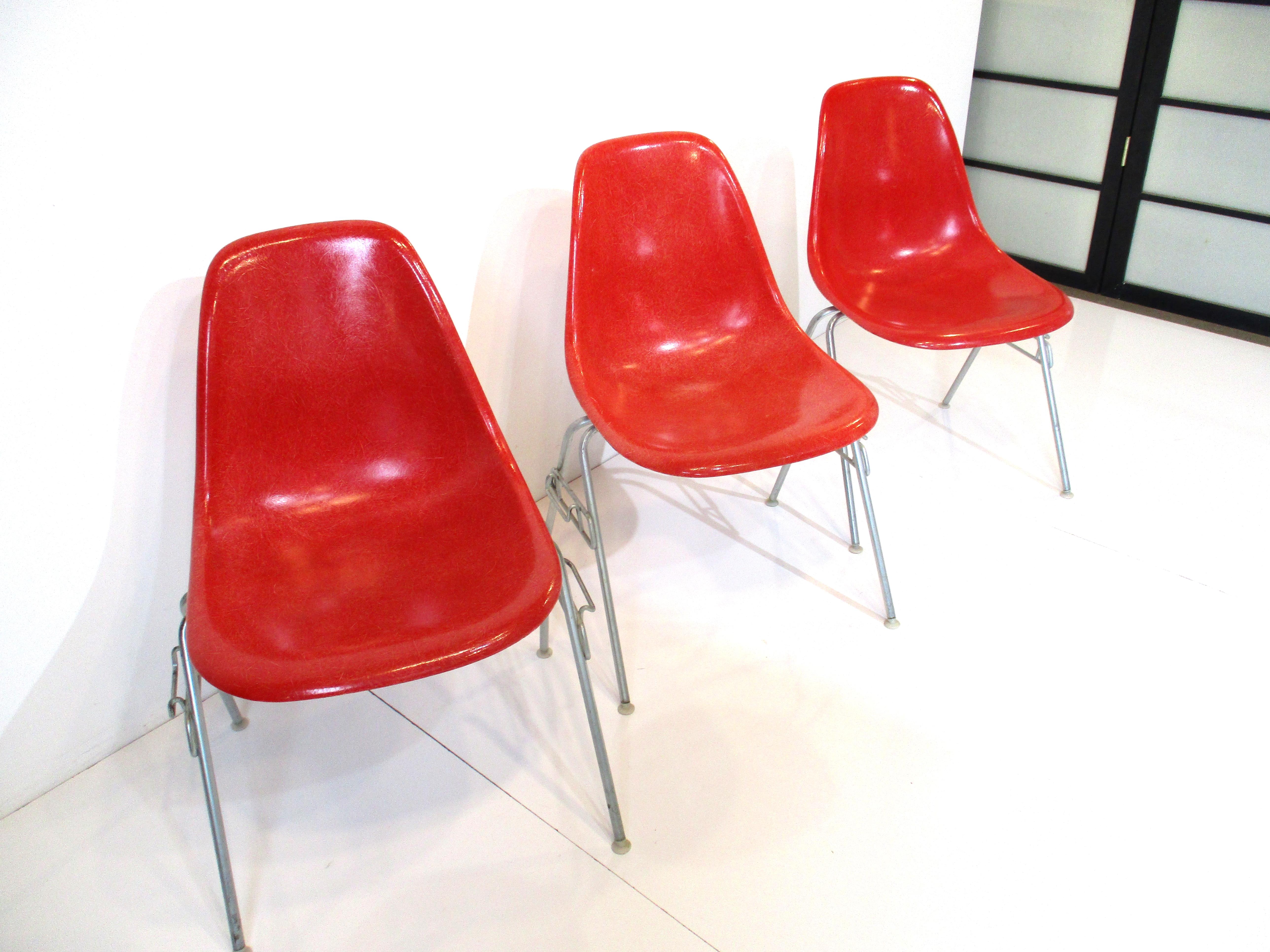 A set of three fiberglass DSS stacking dining chairs with zinc H bases and nylon foot pads . The colors are dark orange , dark red and red with the Herman Miller molded manufactures mark and to the bottoms.