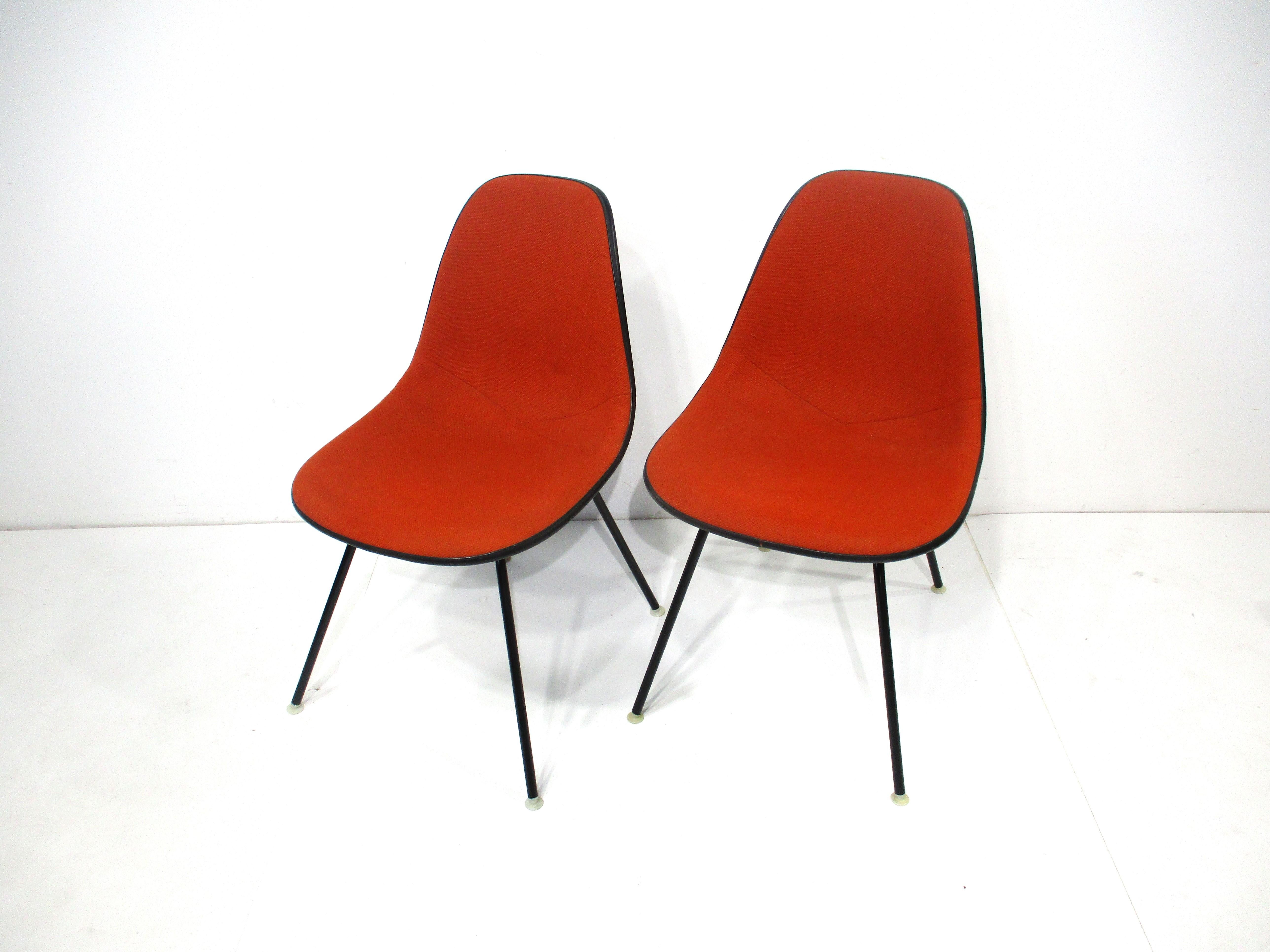 A pair of rust colored upholstered scoop side chairs with black fiberglass shells sitting on satin black H bases . The bases have cream colored nylon foot pads to protect your floors , a classic designed by Ray and Charles Eames for the Herman