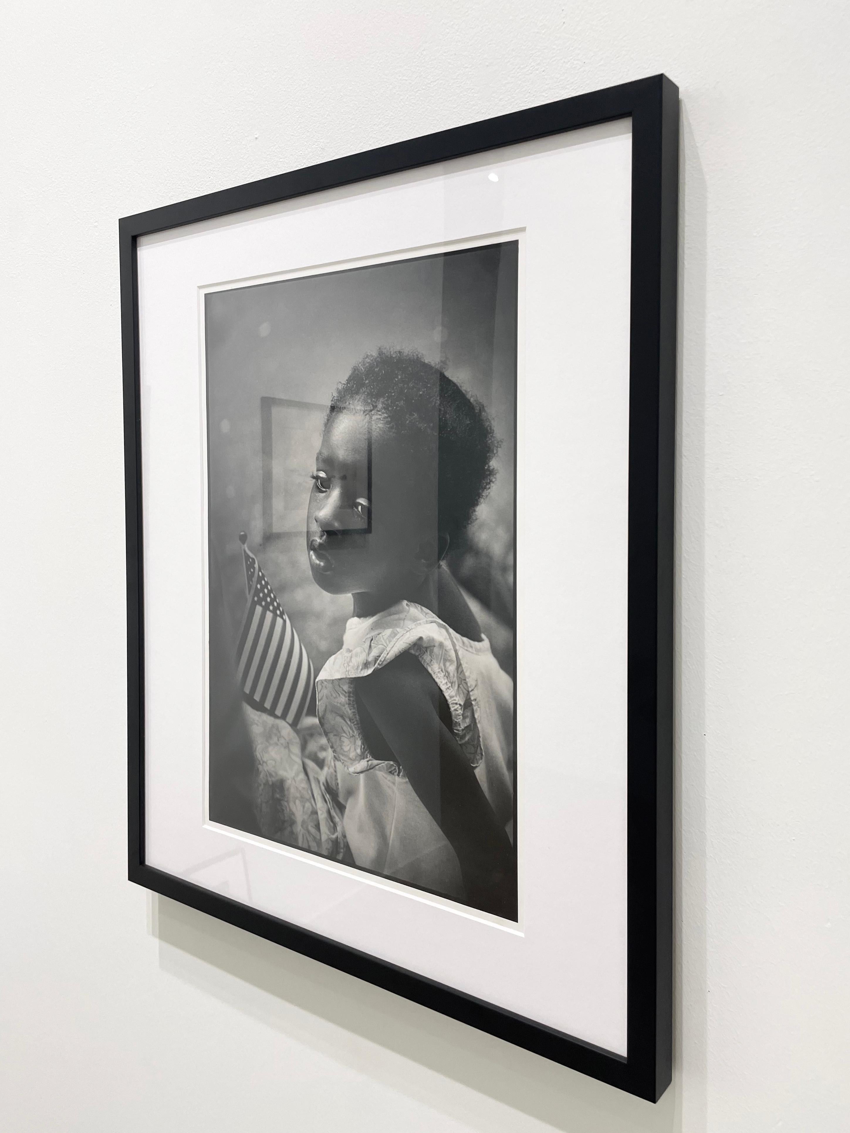 Niña con bandera, de Earlie Hudnall Jr., presenta el retrato de una joven afroamericana que sostiene una pequeña bandera estadounidense. Va vestida de blanco y mira por encima del hombro. Se queda mirando a lo lejos con una expresión inexpresiva,