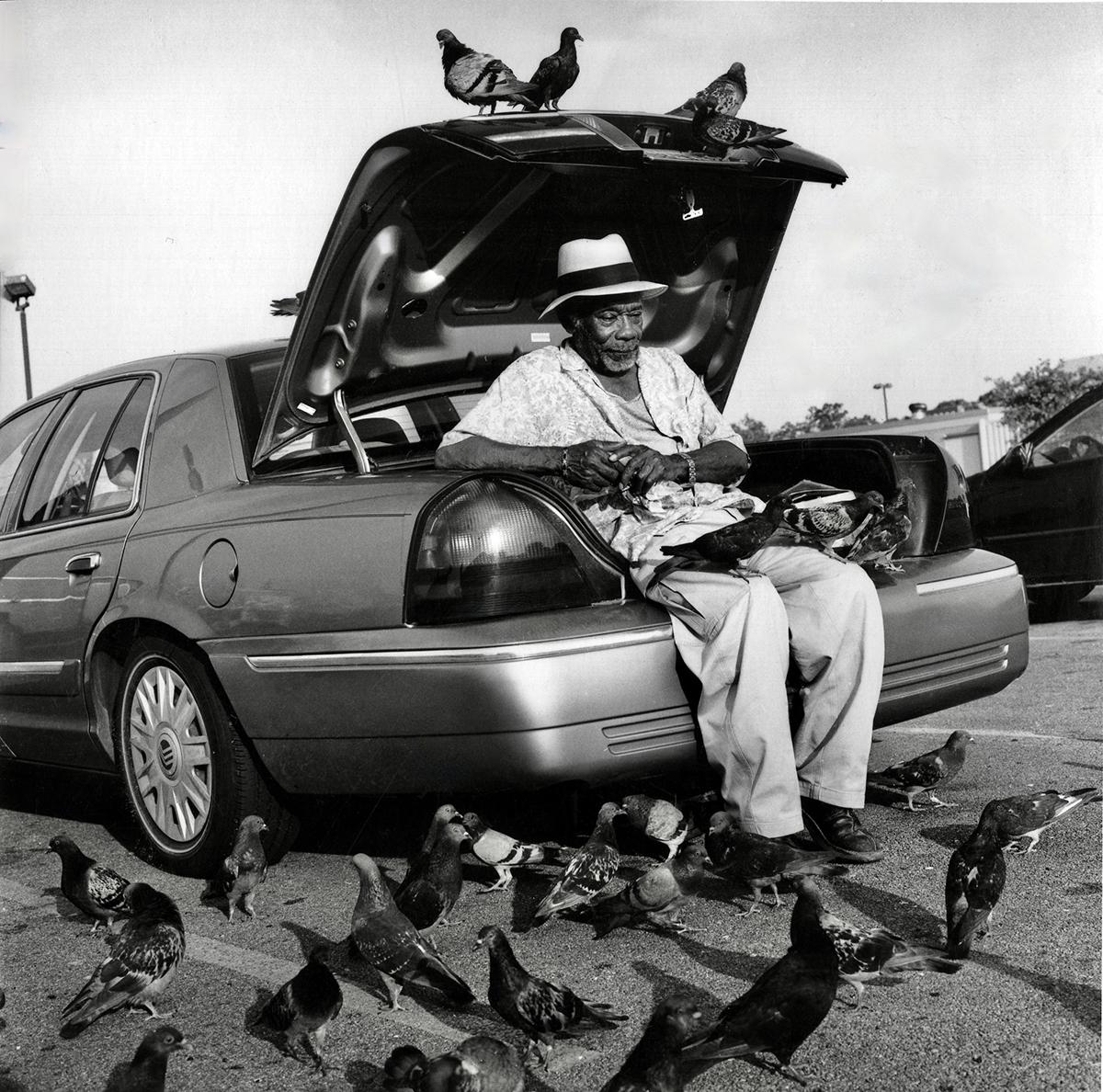 Figurative Photograph Earlie Hudnall Jr. - Homme avec oiseaux - Noir et blanc, homme nourrissant des pigeons, voiture, parking