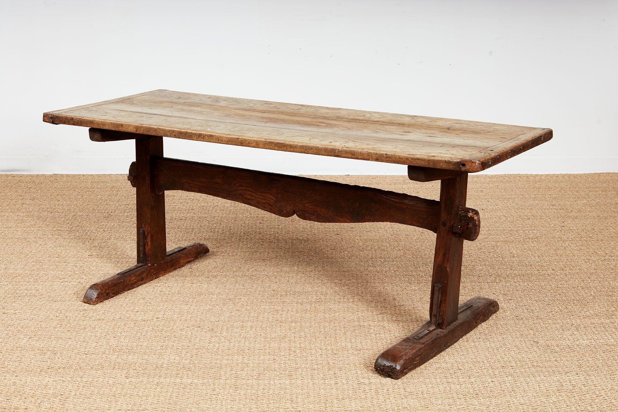 An early 18th century oak tavern table in elm and oak having three plank top with breadboard ends with a scrubbed patina over trestle ends joined by a wide upright stretcher with a shaped underside.  One trestle significantly thicker than the other,