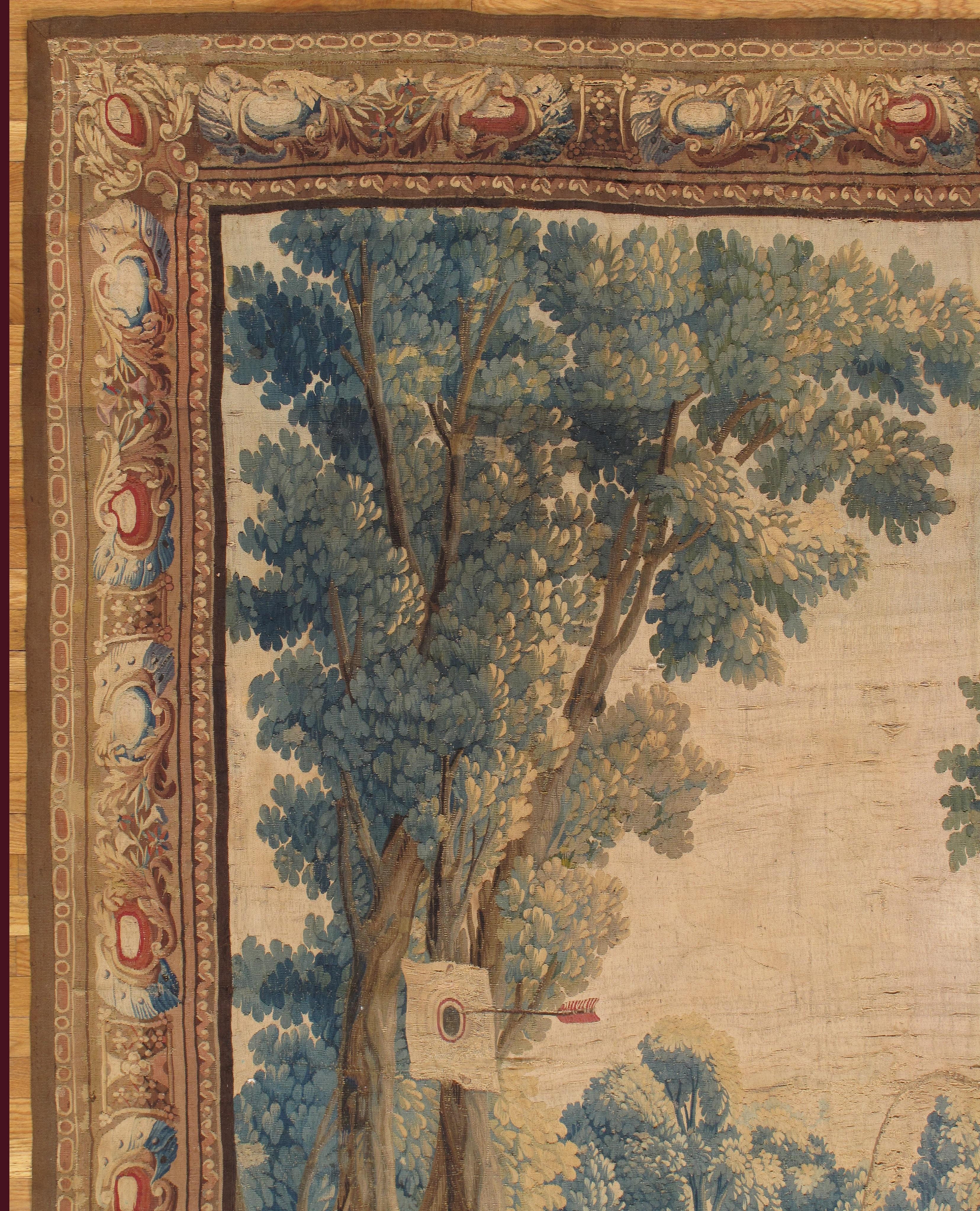 A handwoven Brussels tapestry of dancing peasants in a field with a boy playing jump rope and another with a bow and arrow. 
The foreground shows lush trees and scenery. The border consists of fruits and flowers. 

The colors green, reds, golds, and