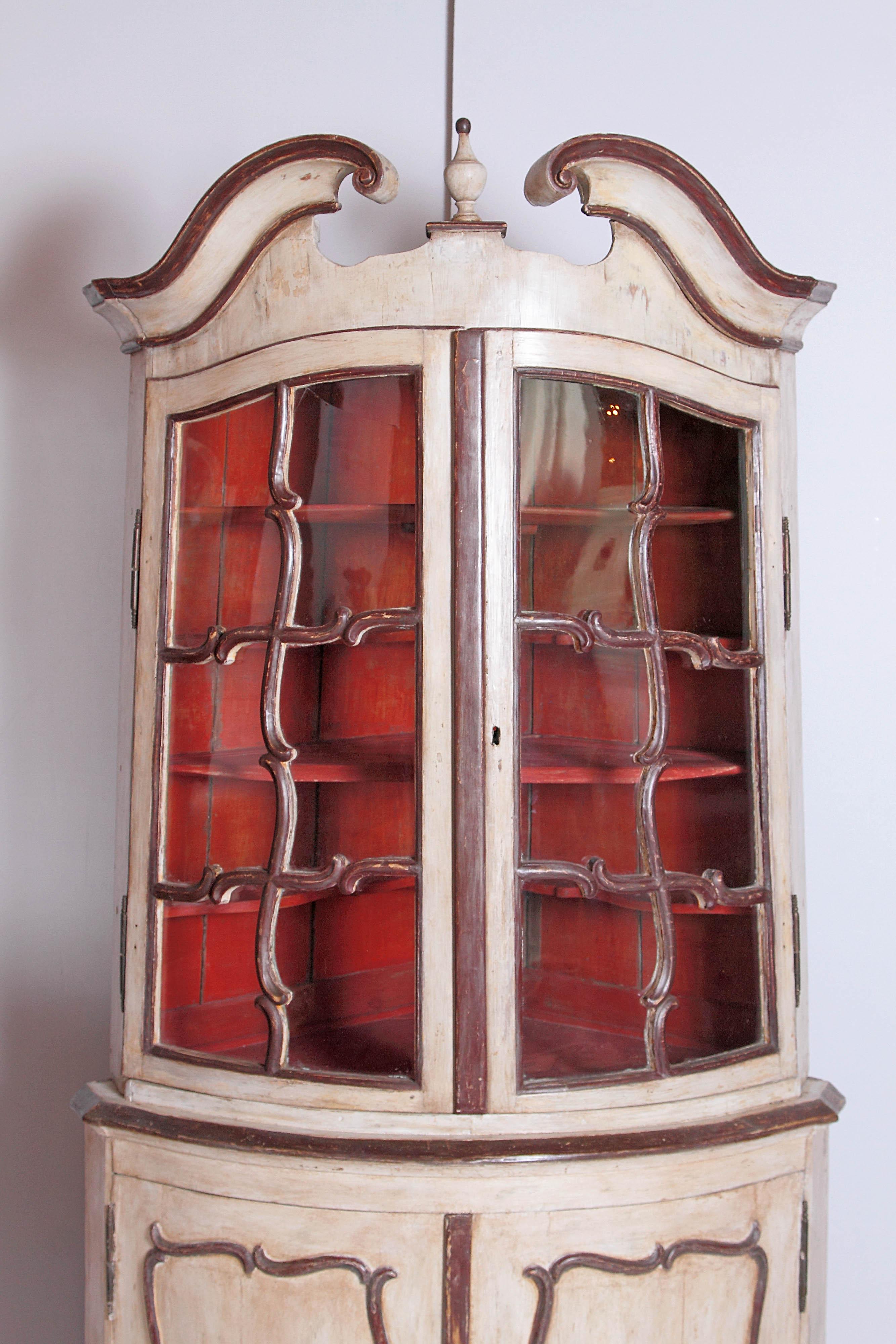 Painted cream with reddish brown trim molding on a bow front corner cabinet with curved glass doors. The cabinet has a split pediment top and urn finial. Behind the glass doors is a painted Asian red interior with four shelves. Below are two doors