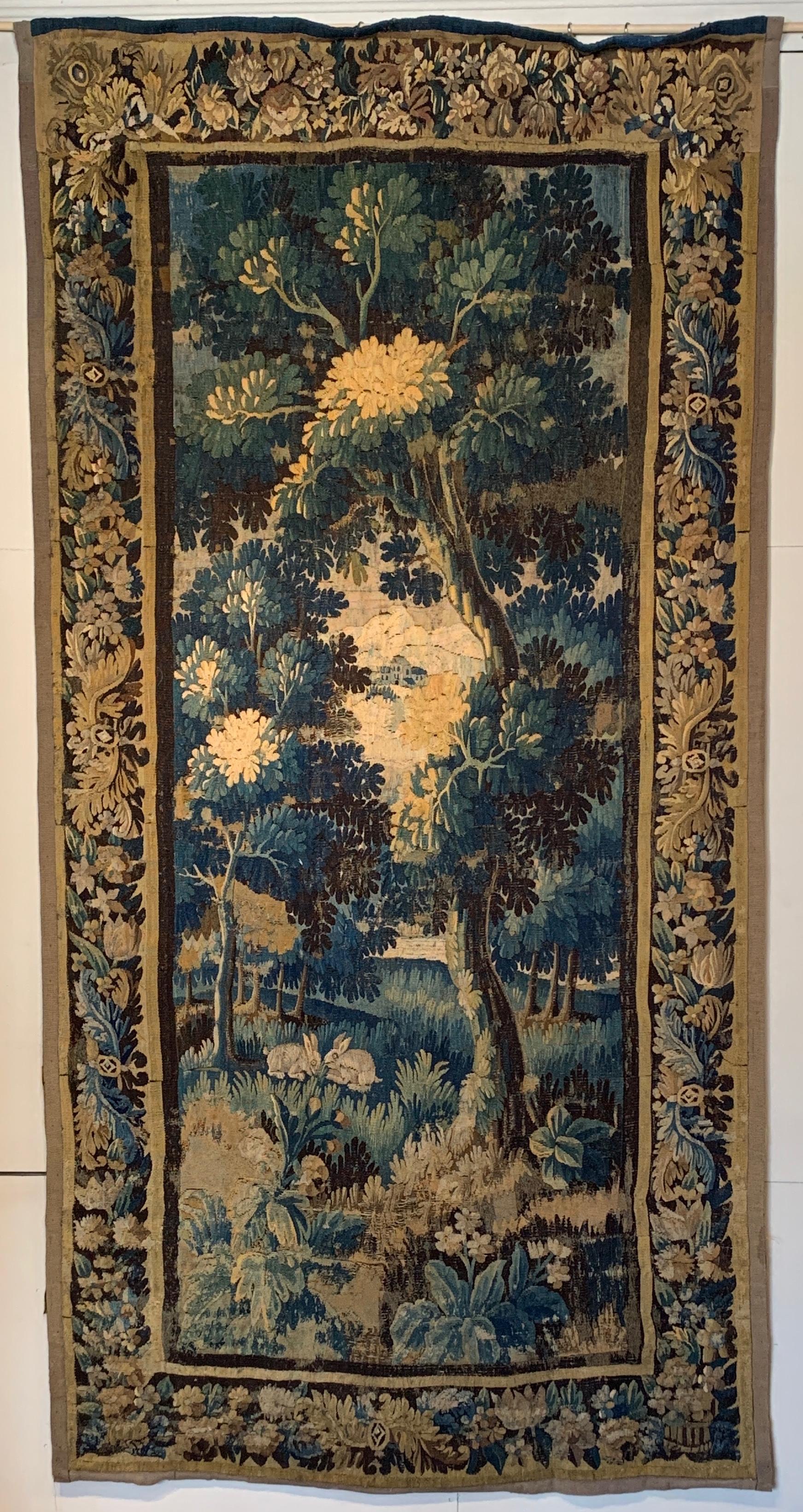 A large early 18th century wool Flemish verdure tapestry depicting an idyllic verdant setting with rabbits and birds, lush foliage and a village in the distance in a traditional palette of soft blues and browns.