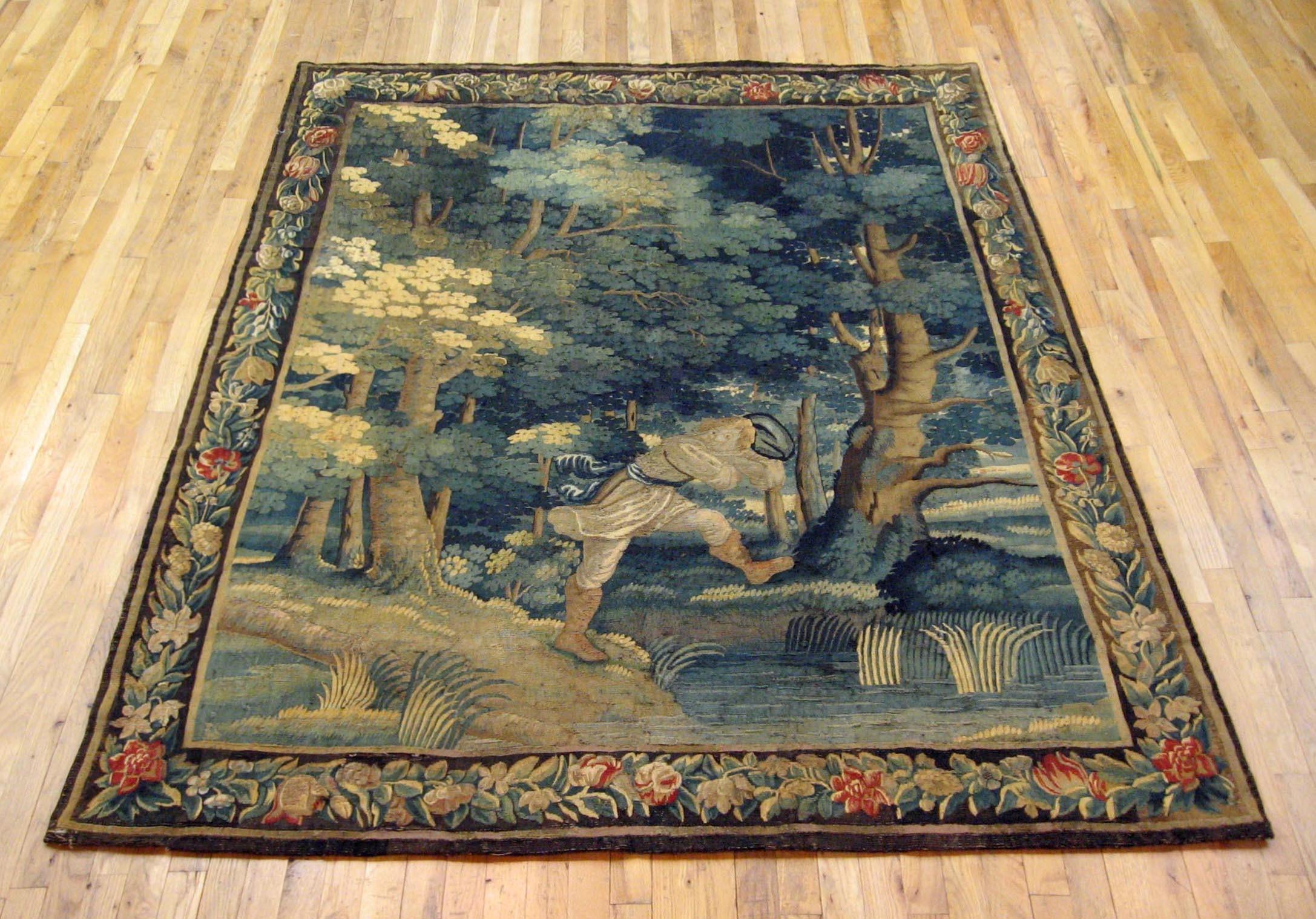 A Flemish verdure rustic tapestry from the early 18th century, circa 1700, depicting a youth a verdant lakeside setting, surrounded by stately trees and other greenery. Enclosed within a scrolling foliate border. Wool with silk inlay. Measures: 7’8”