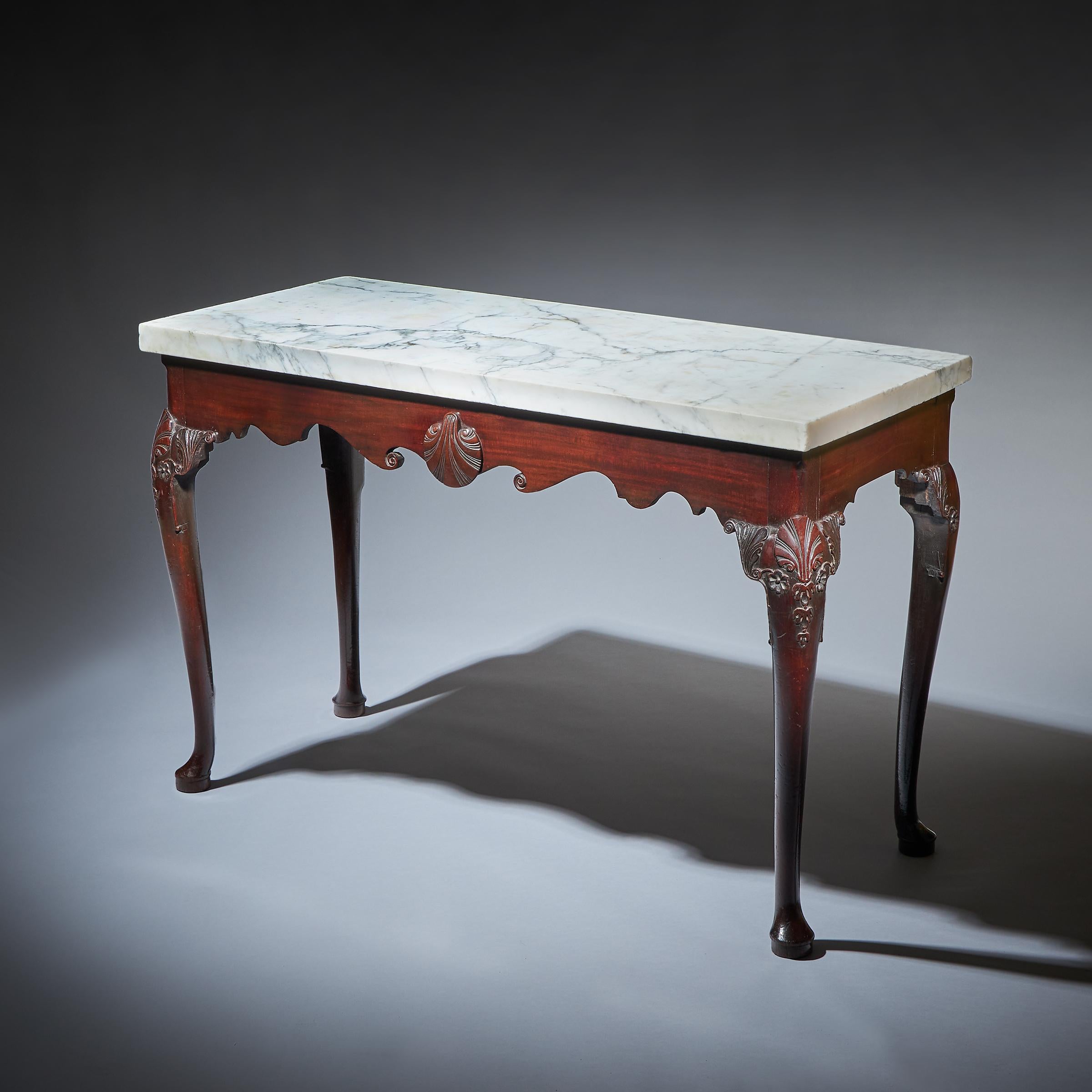 A rare early George I mahogany Irish console table with the original thick-cut statuary marble top over a shaped frieze centred by a carved shell. Raised on eared cabriole legs carved to the knee with a shell, pendant and flower patera leading to