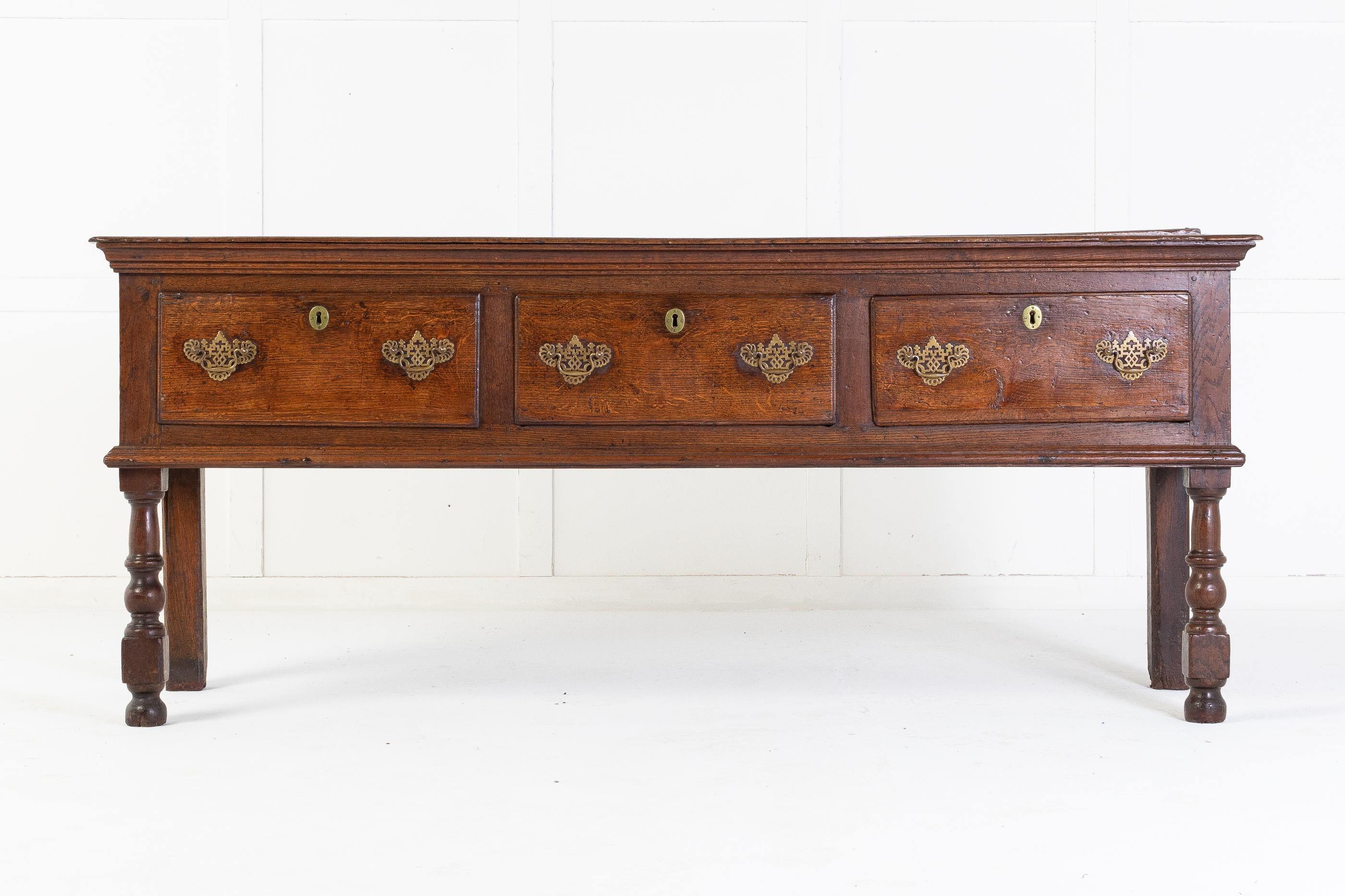 Early 18th century oak dresser base with baluster leg. Having three deep drawers with brass handles and escutcheons.

The top, has two good wide planks of oak and stands on well turned front legs. Deep moulded edges under the top and around the