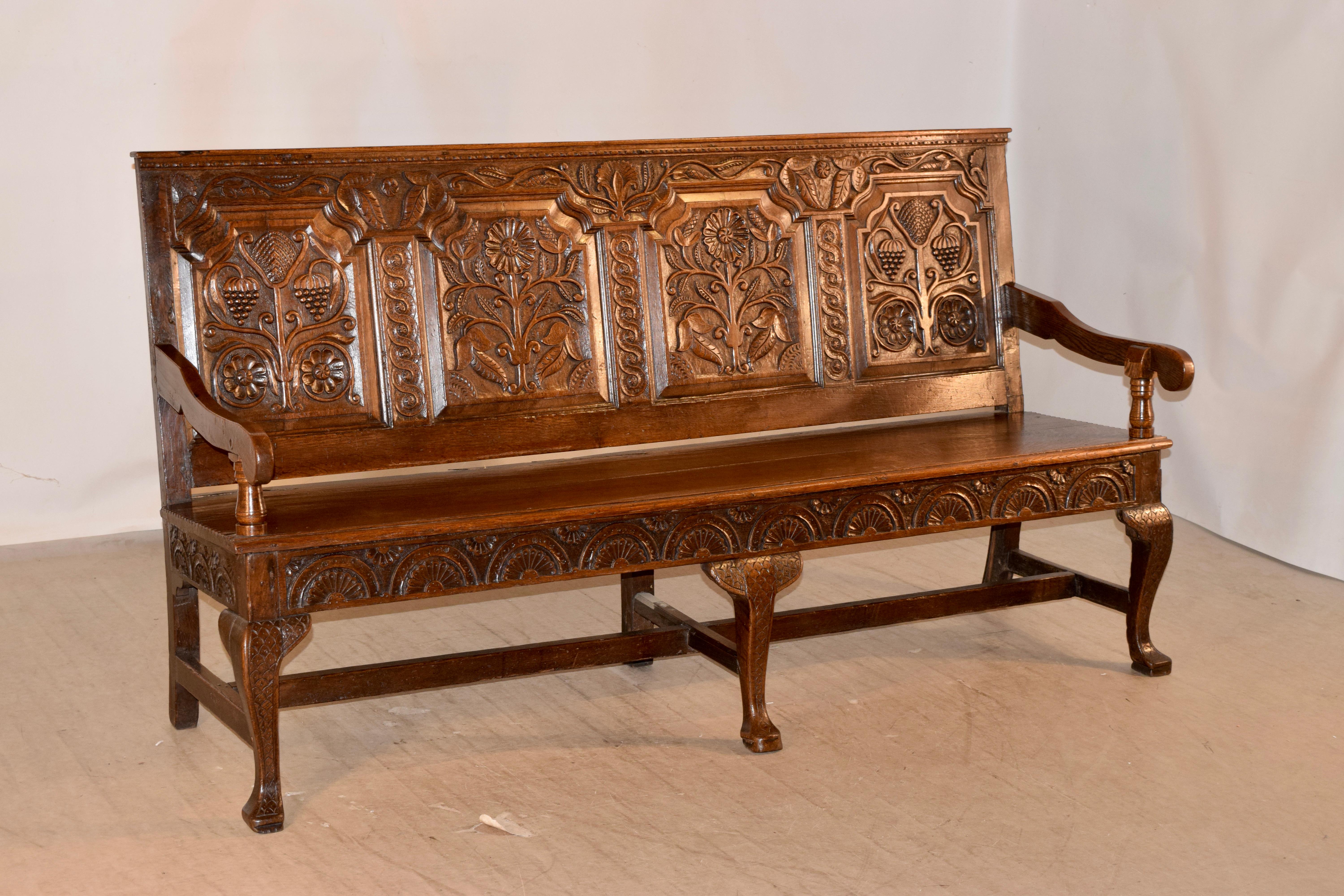 Early 18th century oak bench from England with a highly carved back which has four raised panels, all with floral decoration, flanked with more decorative carvings. The arms are shaped and rest on hand turned supports. The seat is made from two