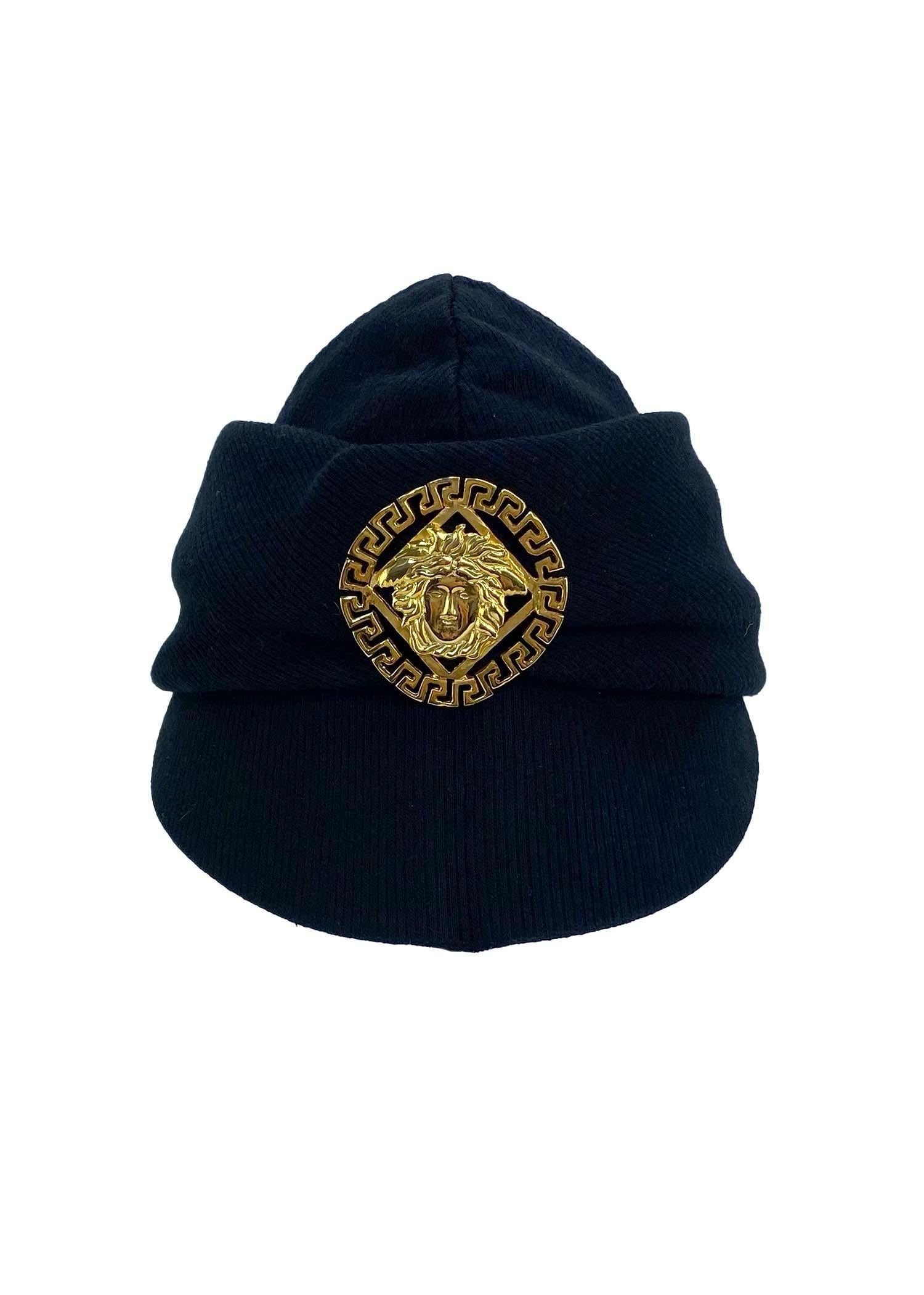 Ti presentiamo un elegante cappello Gianni Versace Couture disegnato da Gianni Versace. Questo cappello con tesa in lana nera, risalente ai primi anni '90, presenta un grande logo Medusa dorato sul davanti. Con una tesa più piccola e un cappello