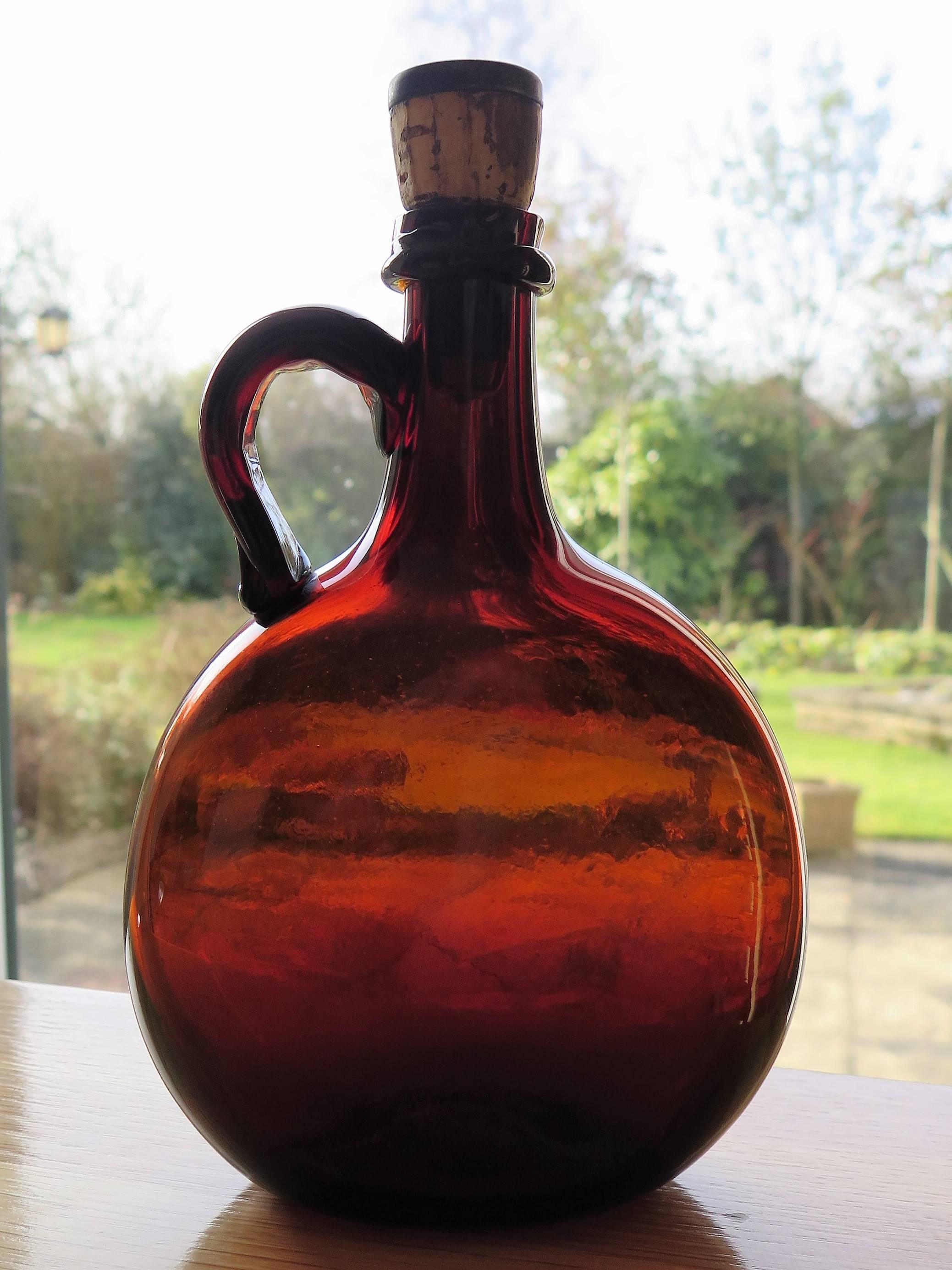 This is a good handblown, amber glass flagon or jug, dating to the early 19th century.

The jug has a circular compressed shape with a loop handle and narrow neck. It is hand blown from amber glass with a rough pontil mark from the blowing process