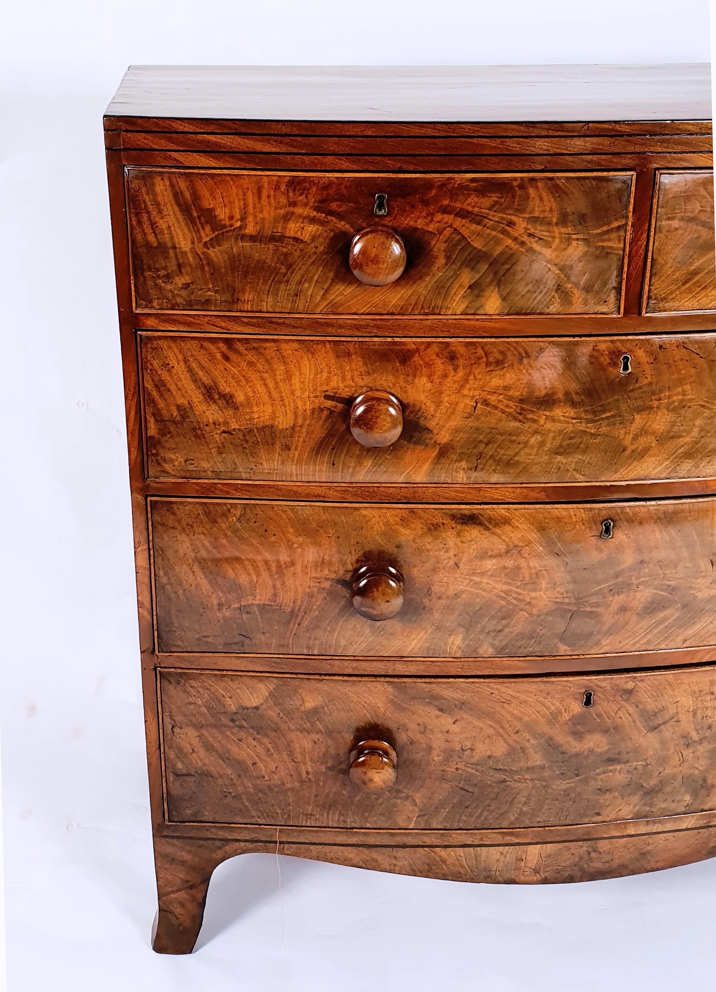 A beautiful and well-proportioned early 19th century English flame mahogany bow fronted chest of drawers that features 2 short over 3 long graduating drawers. The chest has ebony line inlay work and also has the original turned wood handles. It