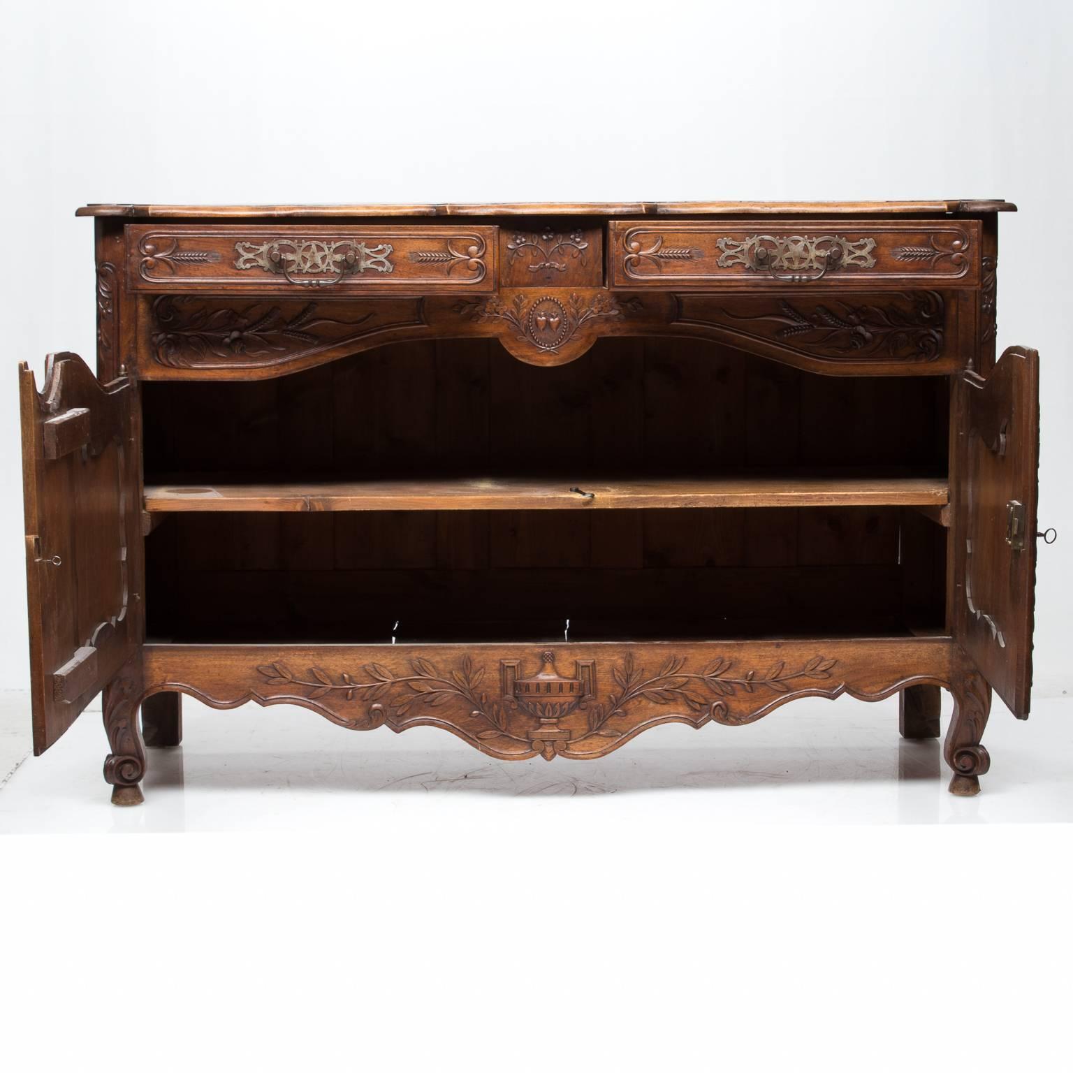An early 19th century walnut French Provincial two-door carved buffet. Very impressive decorative hardware and stout bar hinges. There are two drawers above two doors which open to reveal a single shelf. The top is shaped; the front is cut with