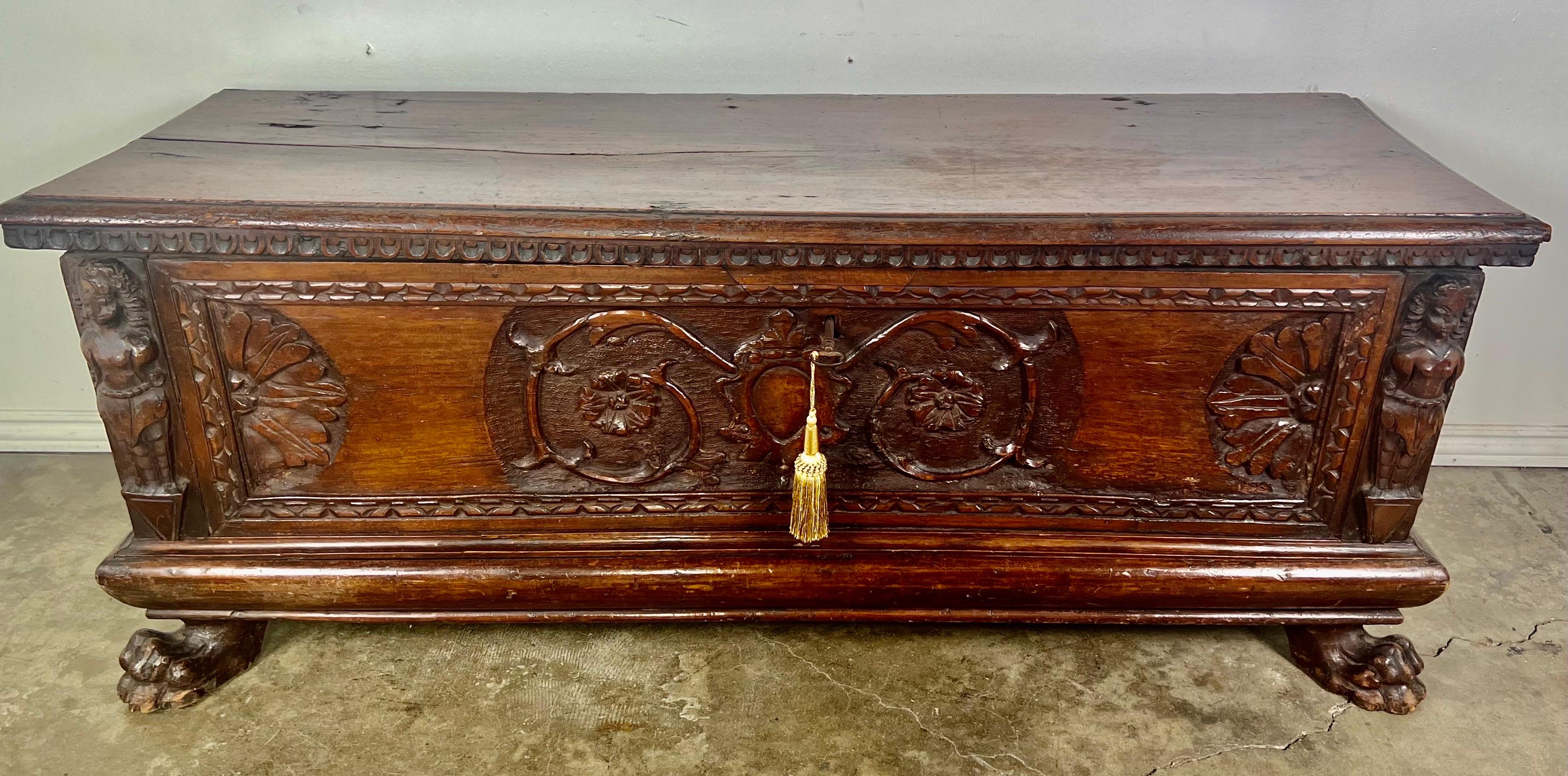 Early 19th century Italian walnut cassone.  It has two carved lion paw feet in the front.  The carving depicts a center cartouche flanked by two scrolled vines ending in center flowers.  There are two figures on either side of the piece.  The walnut