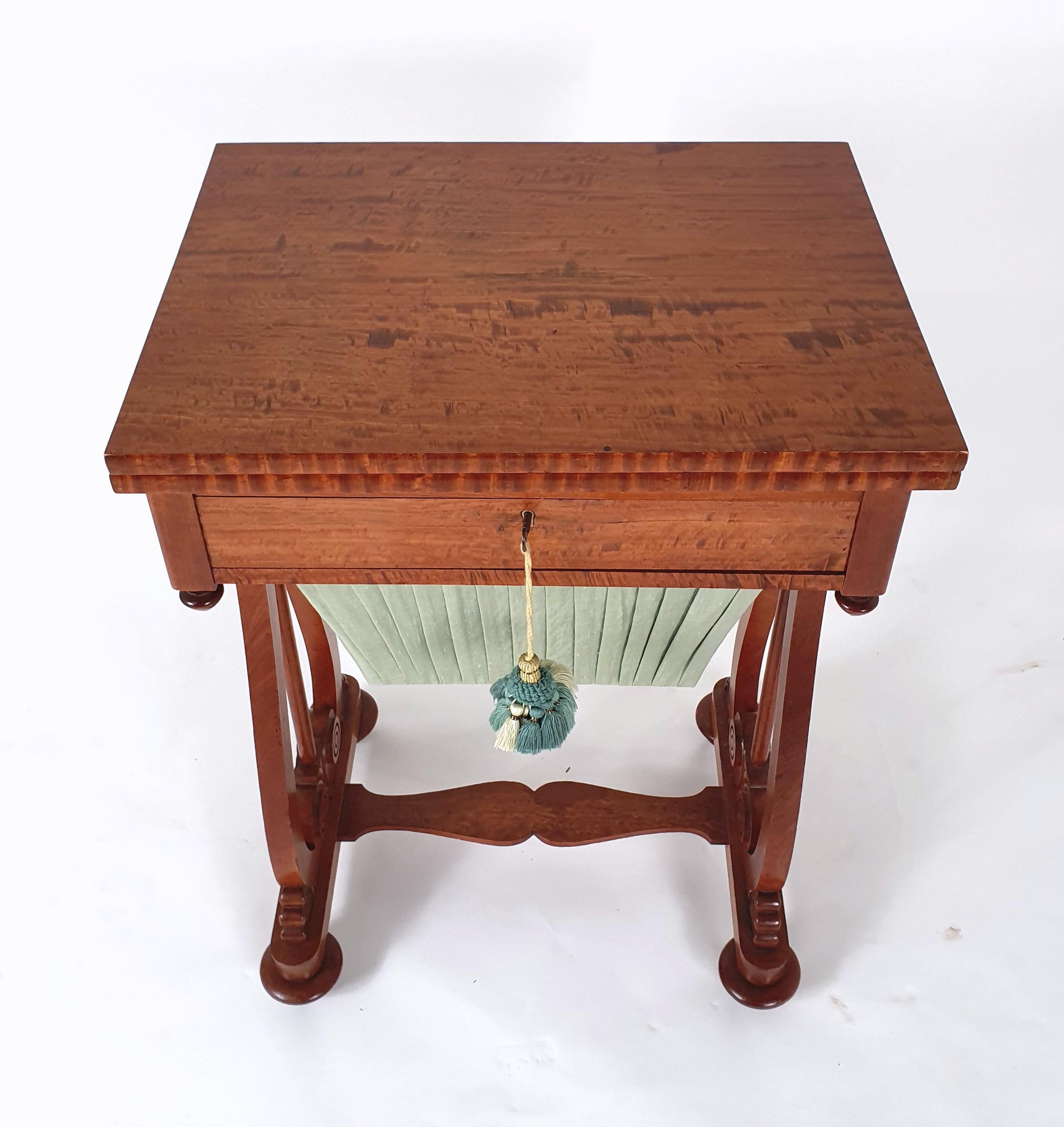 This elegant and very attractive early 19th century English satinwood games/work table features a folding top over a fitted compartmentalized drawer, lined in dark and bright green silk. The table has a work basket, also covered in a pleated pale