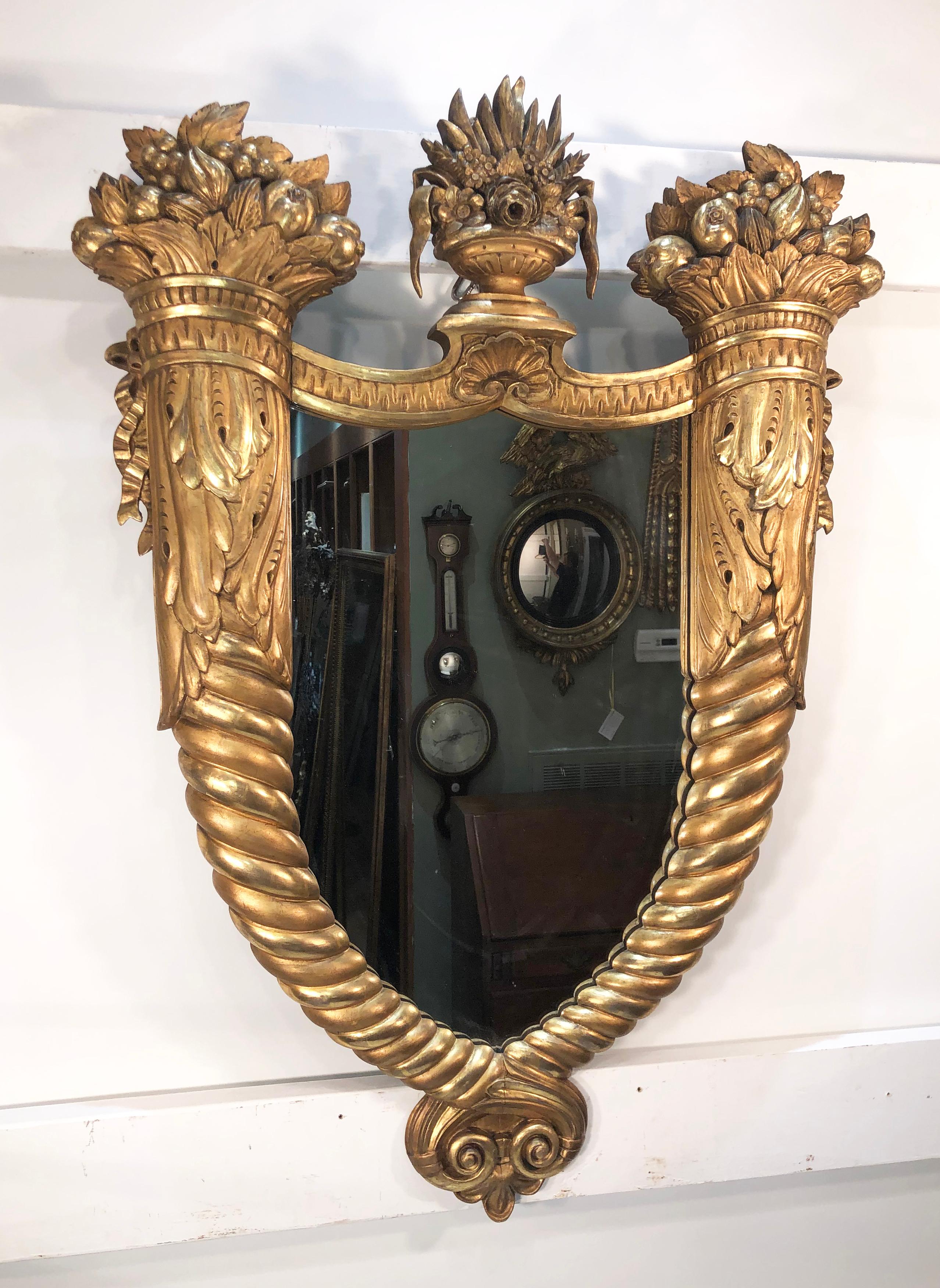 This magnificent carved and gilt wood, circa 1820-1840, Federal Mirror has foliage in a Classical urn finial between opposing Cornucopias. The Cornucopias frame an ovoid shaped mirror. The exceptional frame is carved wood that has gilding in two