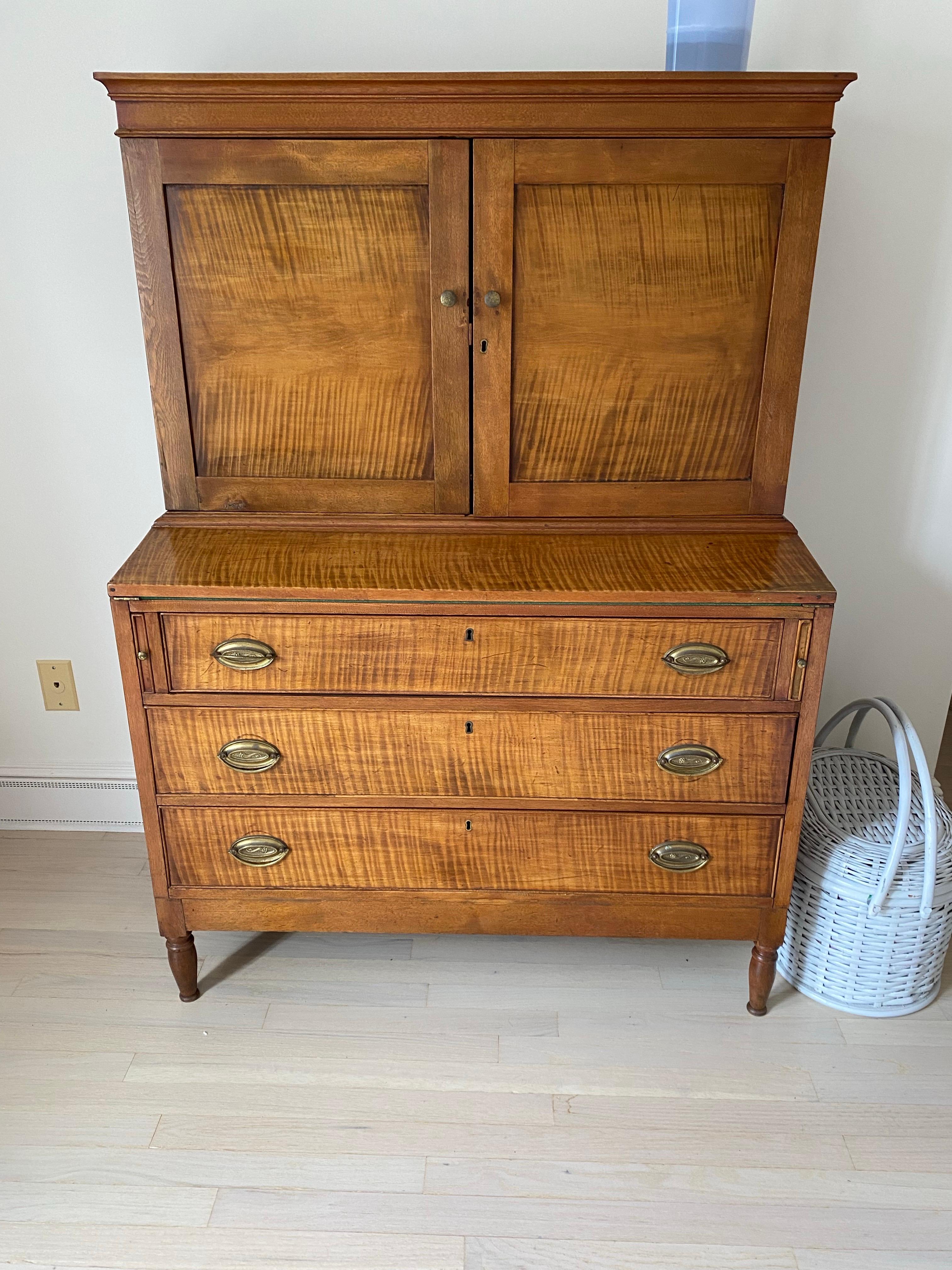 This Early 19th Century American Tigerwood Maple Secretaire. The desk features a drop-front green felted writing surface above a chest of three width-wide drawers below.  A pair of hinged doors reveal open cubbies above three small drawers for