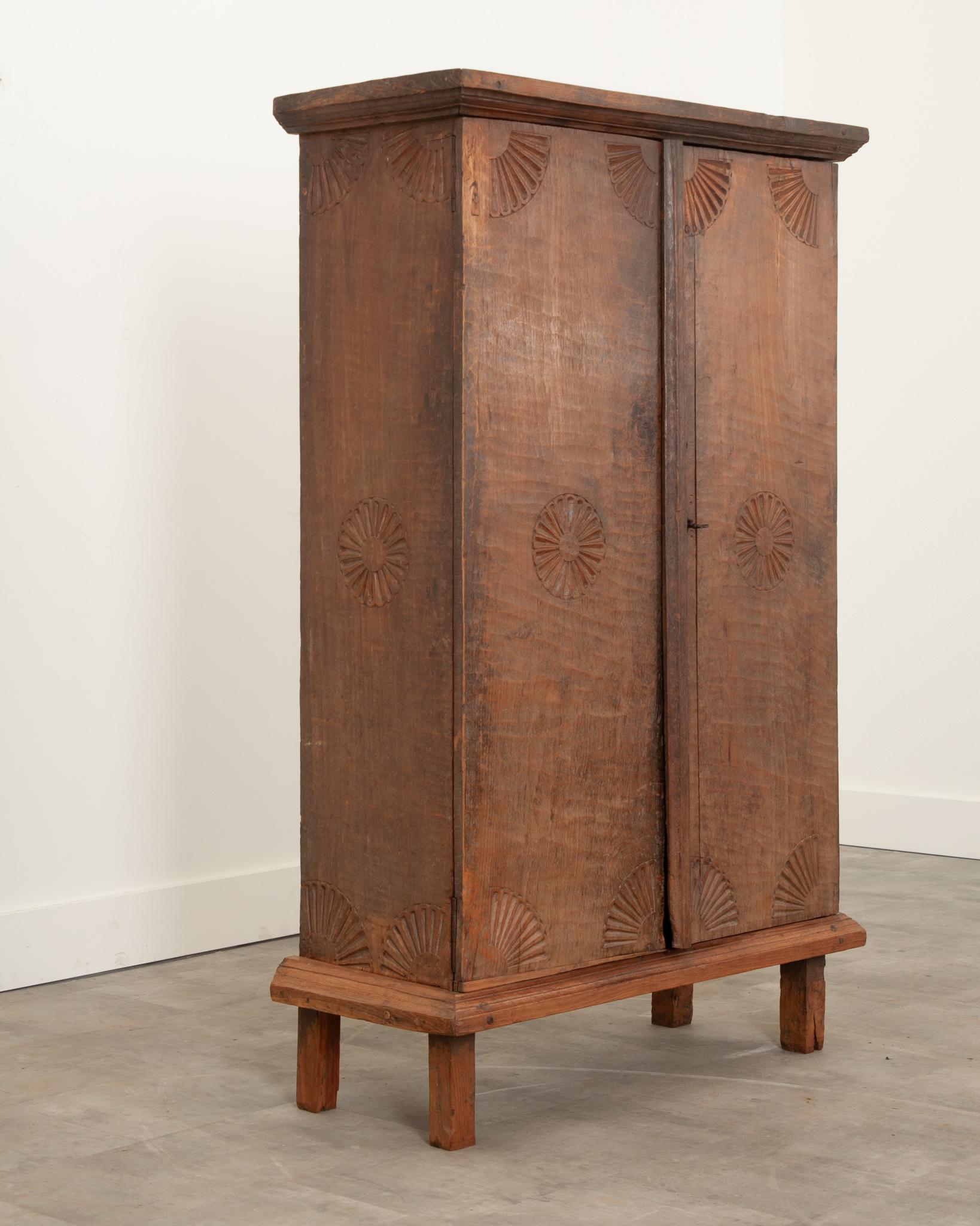 A primitive teak cabinet from British Colonial India. The cabinet has been totally carved and planed by hand. It features a repeated carved sunburst motif found on all panels. The interior has two fixed shelves and can be secured with the cabinet’s
