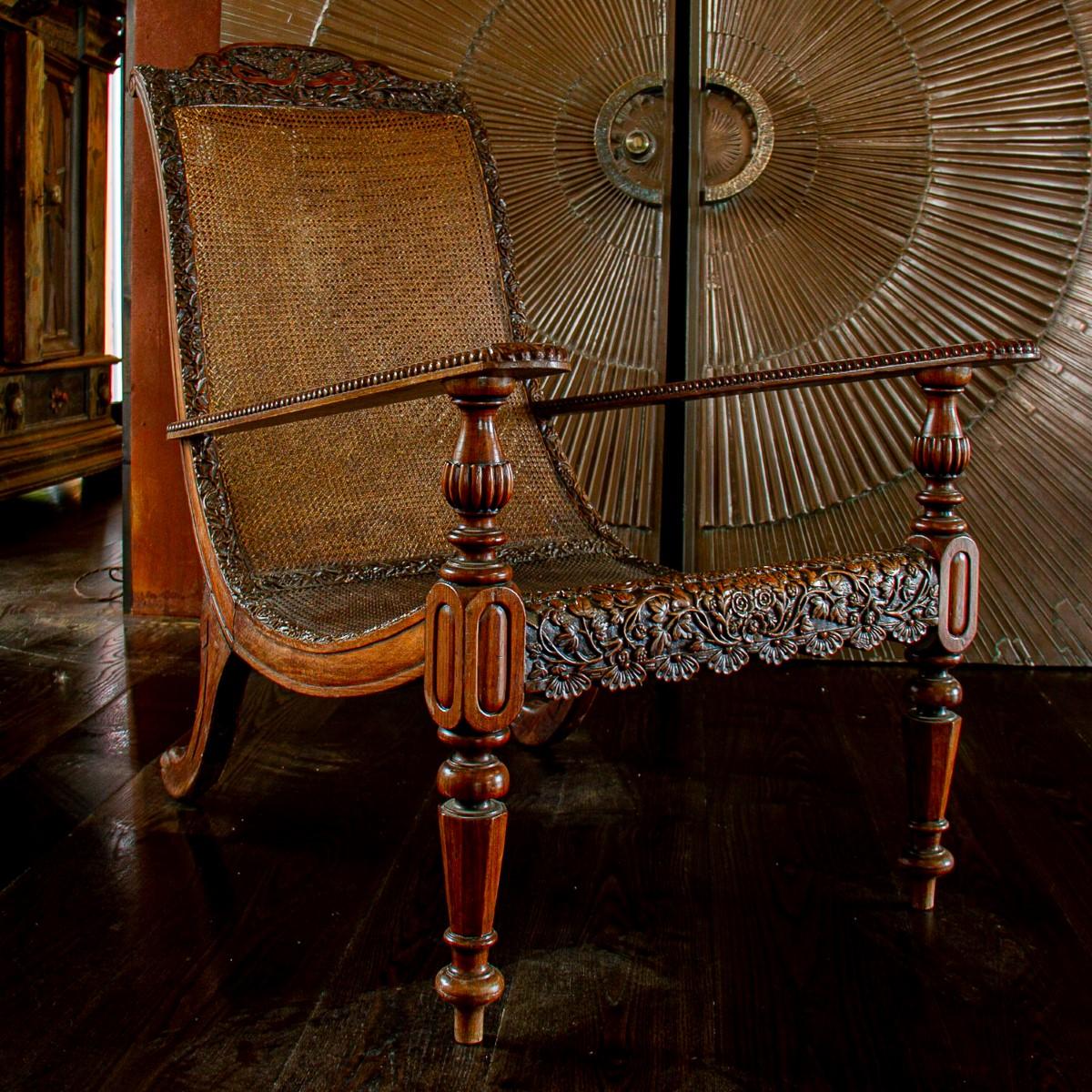 A 19th century Anglo-Indian padouk wood armchair with carved eagle crest amongst foliage. The rest of the frame is heavily carved with flowers and leaves around a rattan seat and backrest. The arms are removable from the turned padouk arm supports