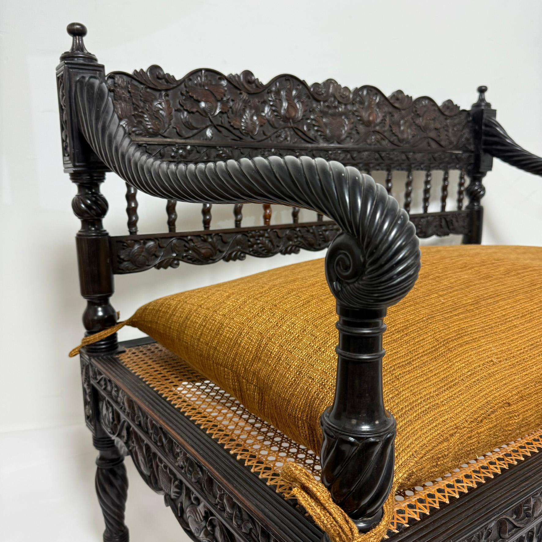 An early 19th century Anglo Indian caned seat settee (with down filled cushion) in extensively carved solid ebony from the British colony of Ceylon (Sri Lanka today). The exceptional craftsmanship of these Indian makers was heavily influenced by