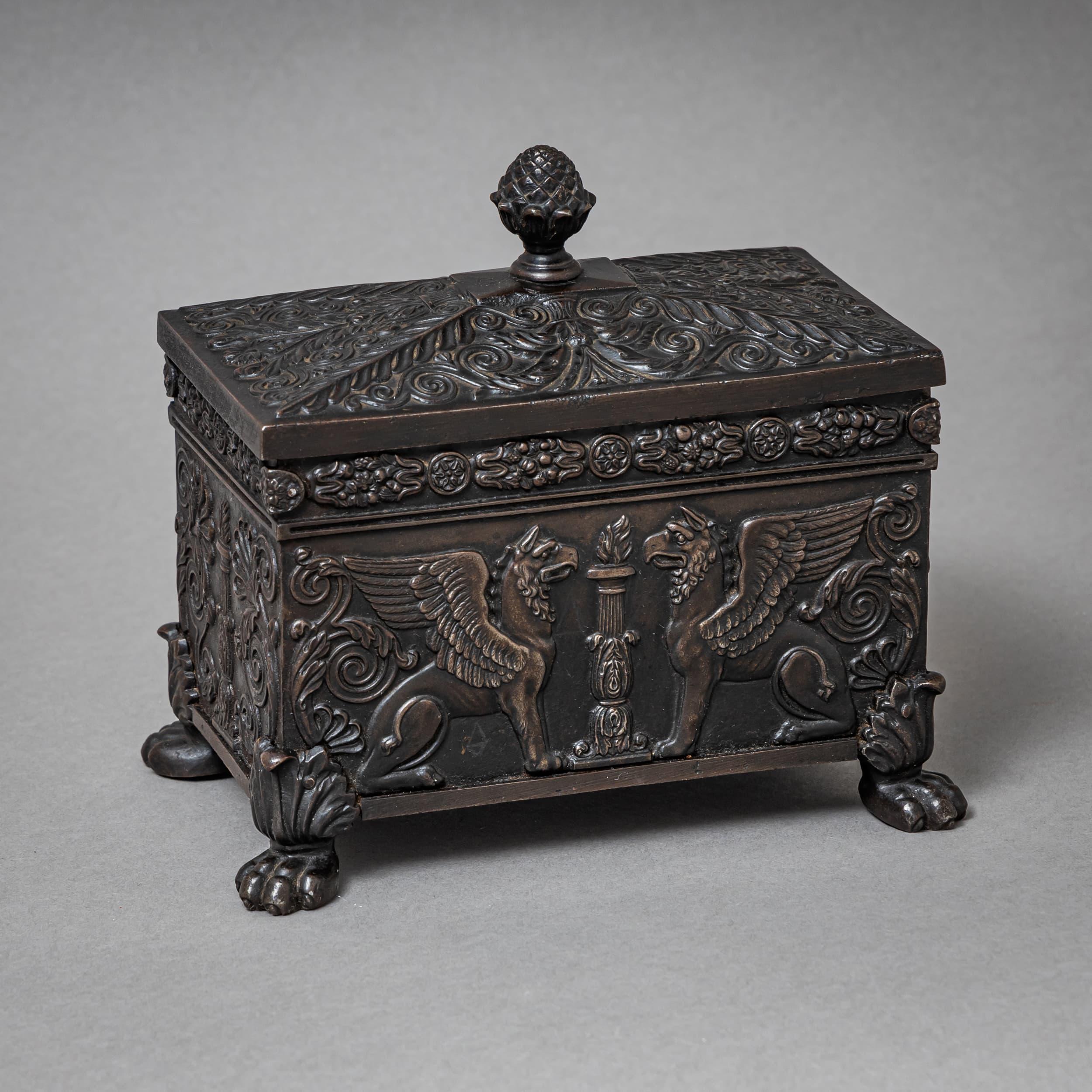 Rectangular Berlin iron tobacco box dating to the early 19th century, richly adorned with intricate ornamental detail. The pyramidal lid is topped by a finial shaped like a pinecone. Resting on sculpted lion paw feet, the casket is decorated with