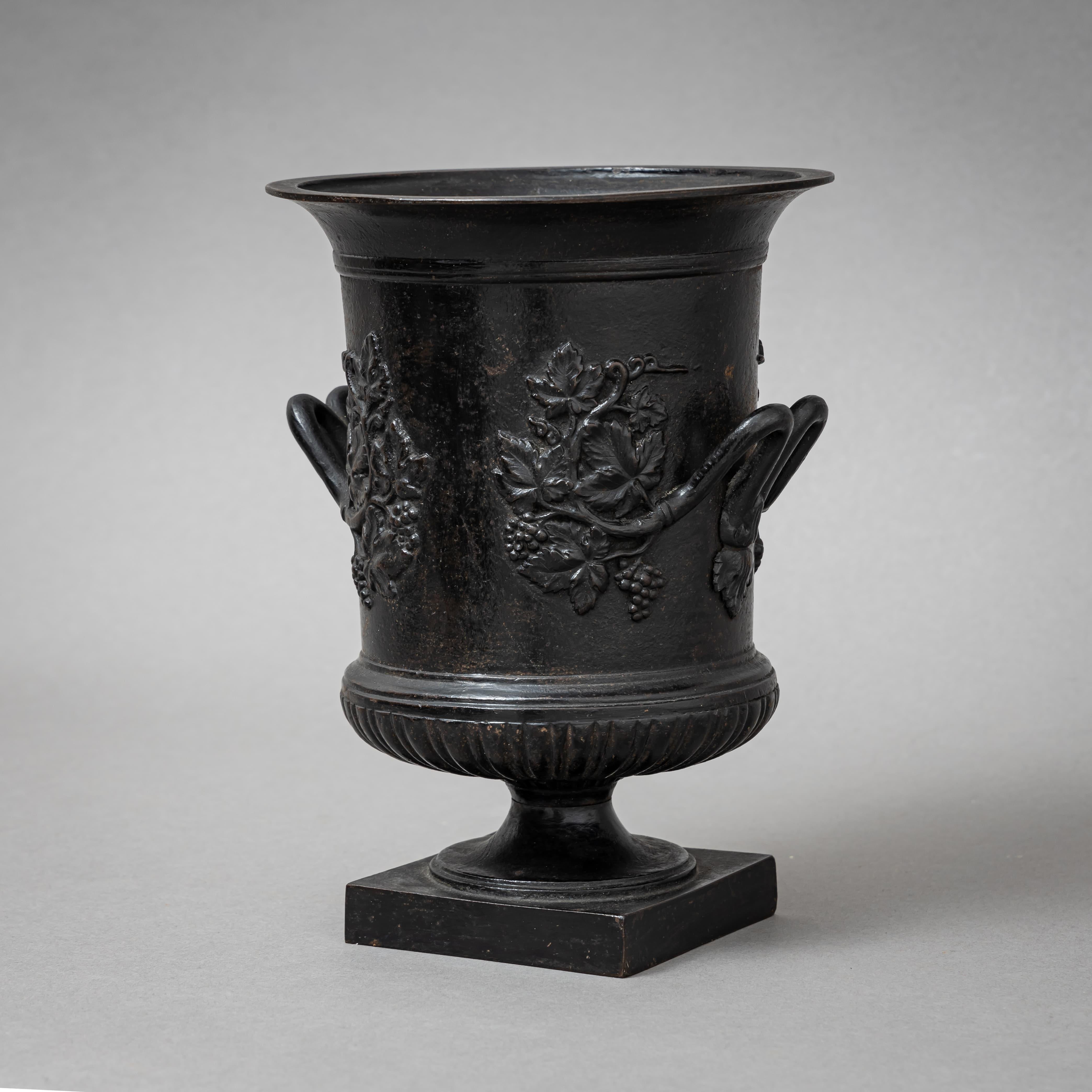Vase cylindrique en fer de Berlin avec un bord évasé et des poignées latérales. Le corps est décoré de motifs de vigne et de raisin en relief. La tige en forme de balustre repose sur une base carrée.