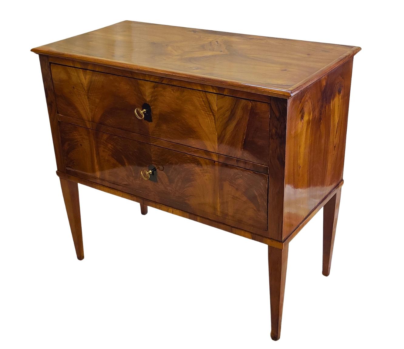 Questa rara commode a due cassetti è stata realizzata nella Germania meridionale intorno al 1790-1810, nel periodo di transizione tra il tardo classicismo e il Biedermeier. L'elegante pezzo è impiallacciato in noce, splendidamente abbinato a tutti i