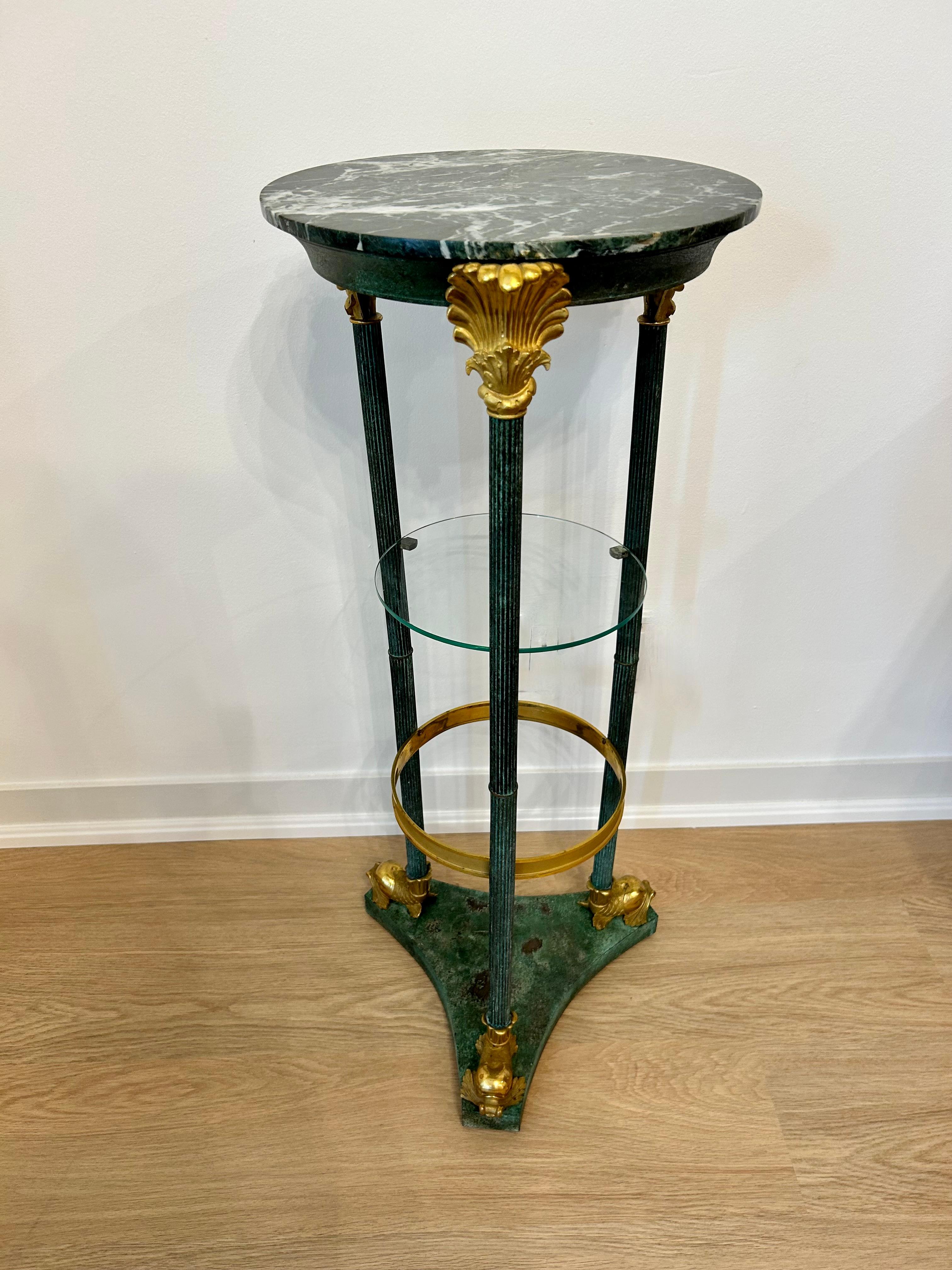 19th Century patinated metal pedestal table with three legs and a triangular base in verdigris finish. Gilt  gilt bronze dolphin feet, floral capitals and an interior ring. The top is green marble, and there is a lower glass shelf. Both of these may