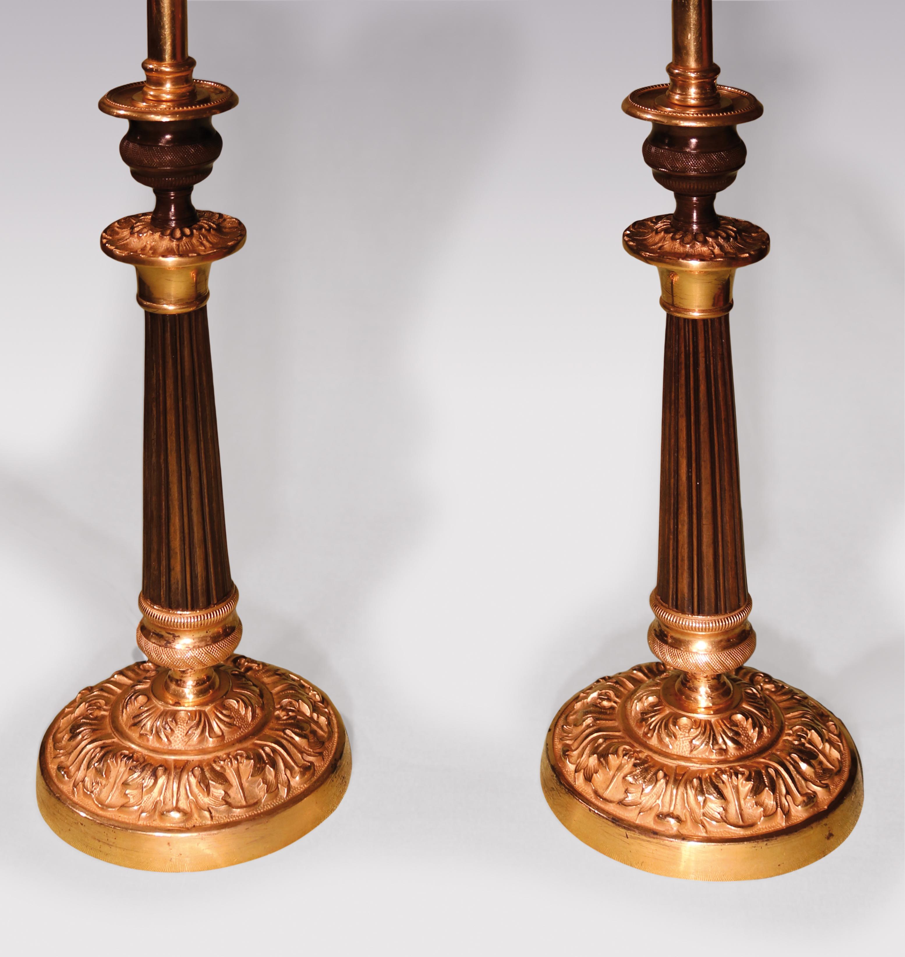 A pair of early 19th century bronze and ormolu candlesticks having engine-turned vase-shaped nozzles above reeded tapering stems ending on acanthus decorated circular bases. (Now converted to lamps) Height with shades: 20.50 