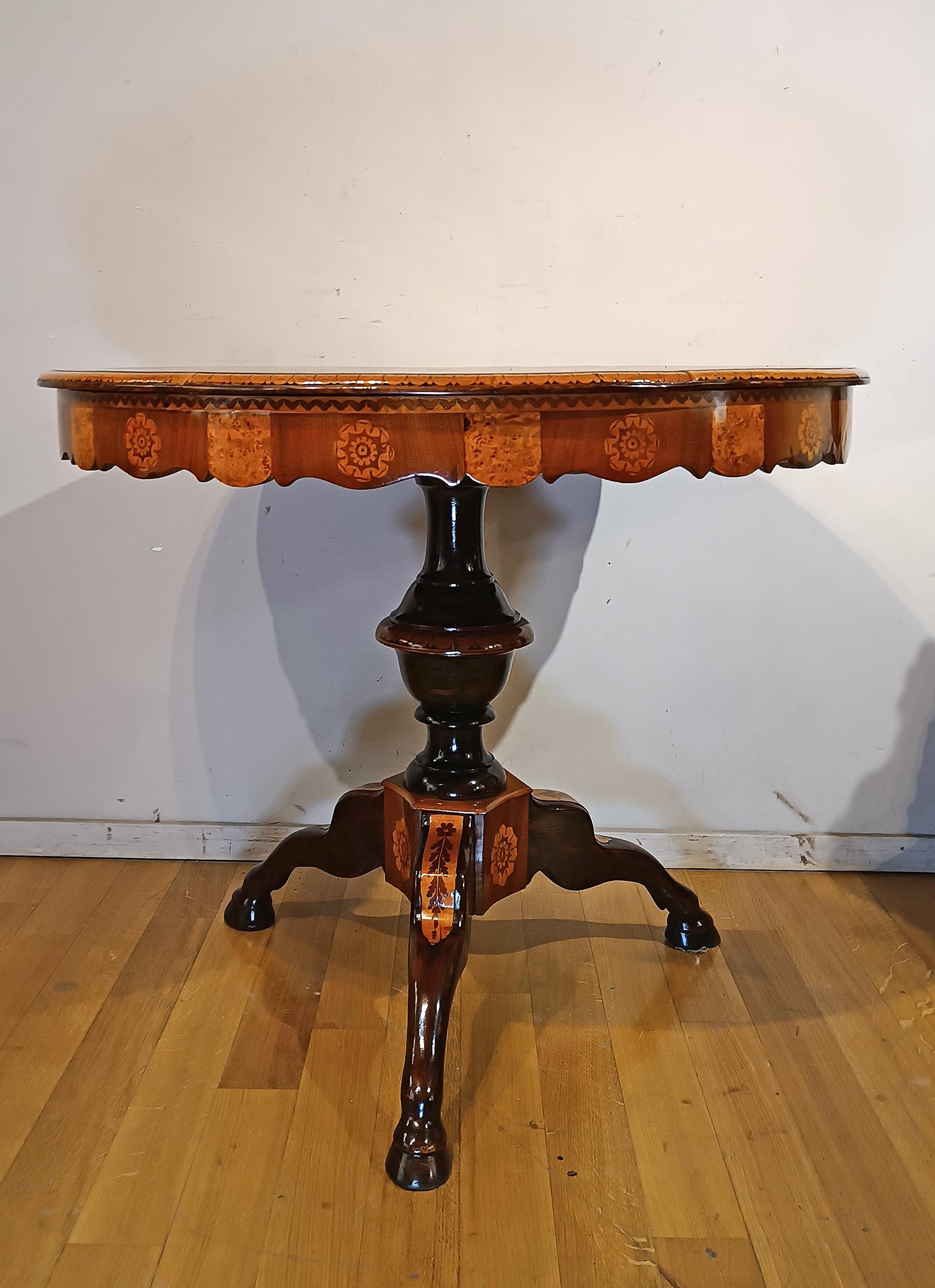Elegant circular center table in carved walnut, embellished with inlays in walnut feather, walnut and fruit woods. The structure has a curved band and a molded central shaft, which ends with three-legged feral feet with a plinth. The table stands