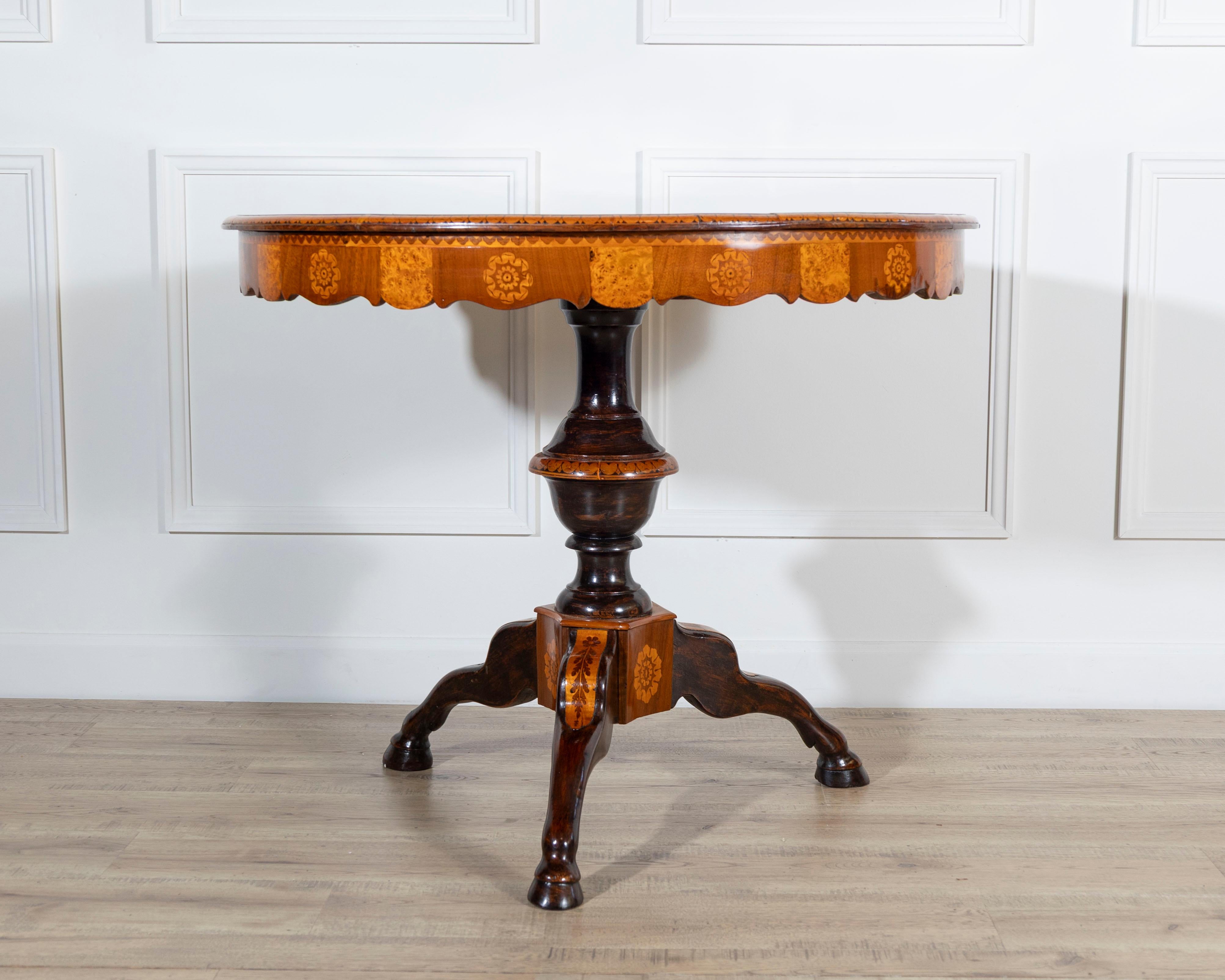 Elegant circular center table in carved walnut, embellished with inlays in walnut feather, walnut and fruit woods. The structure has a curved band and a molded central shaft, which ends with three-legged feral feet with a plinth. The table stands