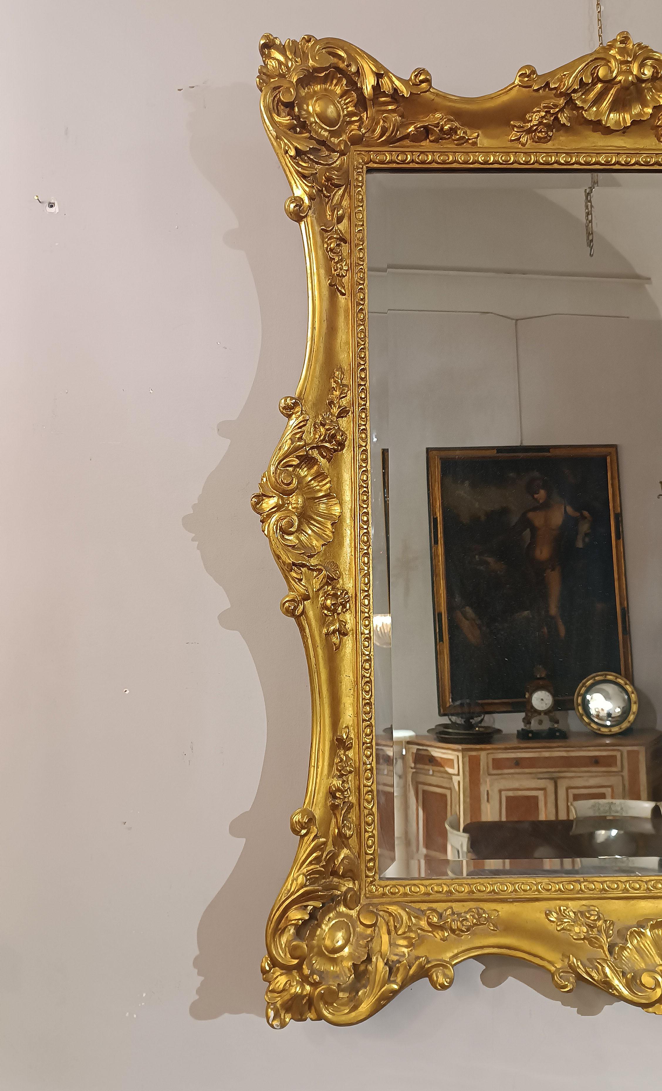 Magnificent and elegant mirror in carved and gilded wood with pure gold leaf. The frame, with a curved shape, is enriched with geometric decorations and vegetal scrolls that give a touch of class and sophistication. The mirror glass is original and