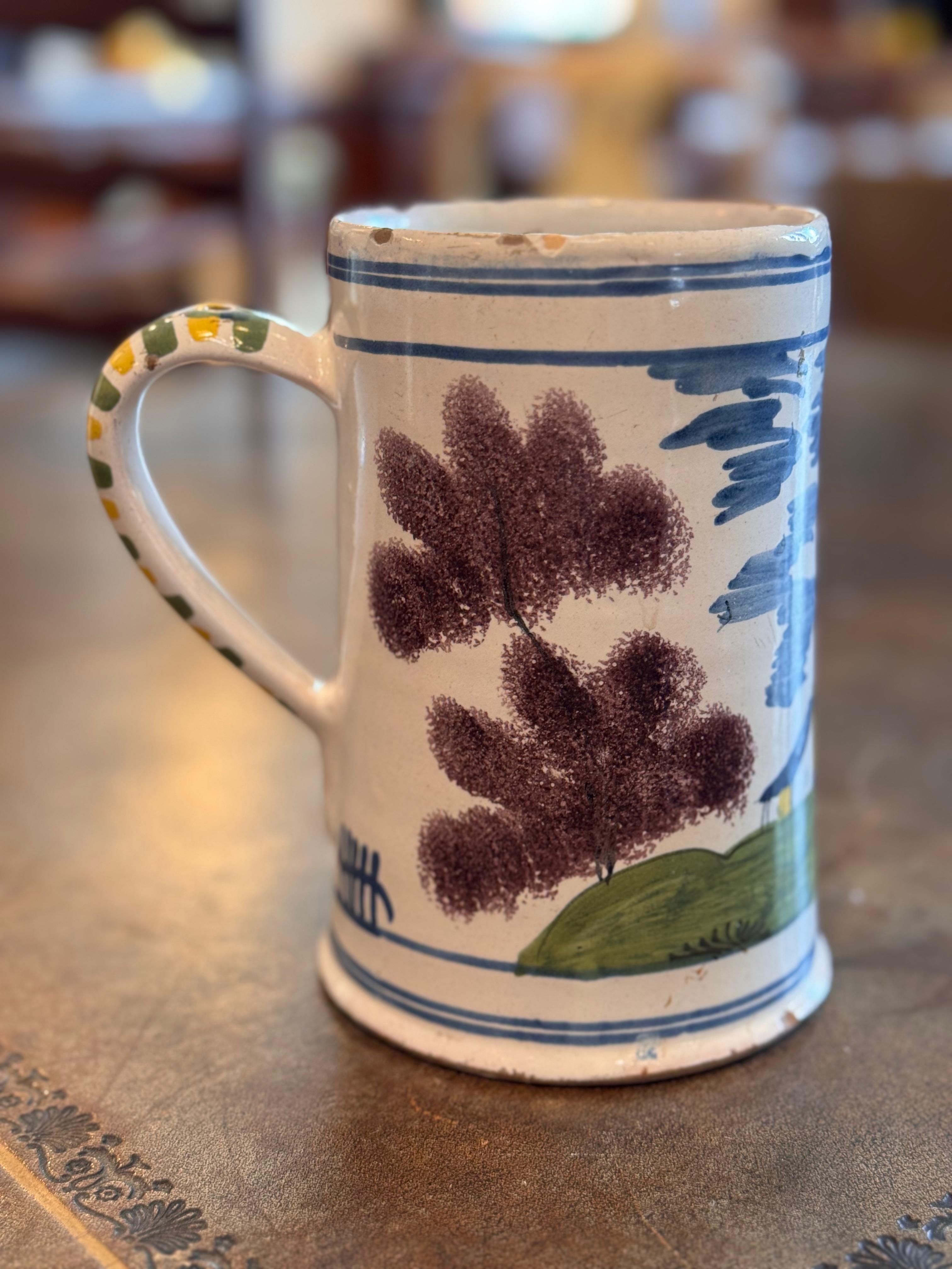 Une chope de Delft peinte à la main et représentant un paysage hollandais traditionnel. La forme cylindrique est décorée d'un moulin à vent, d'arbres et de chalets dans les tons classiques bleu, vert et jaune. La poignée est rehaussée de détails
