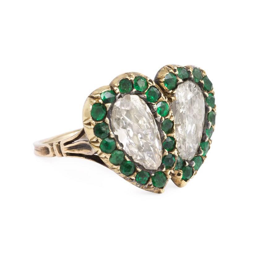 An antique gemset close-backed ring designed as two overlapping diamond hearts with emerald surrounds, in sterling silver and 14k gold.  Diamonds measure approximately 9.3 x 5.3mm and 9.6 x 5.7mm, respectively.  Top of ring measures 9/16 x 3/4 inches