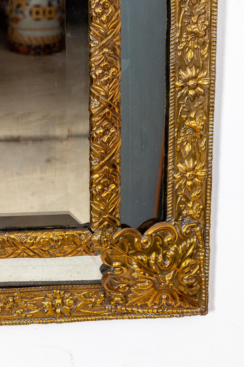 Circa early 19th century Dutch metal framed mirror in a repoussé finish with floral motifs throughout and a central beveled mirror plate. The mirror is capped with a large triangular shaped pediment detailed with beaded trim and scrolled acanthus