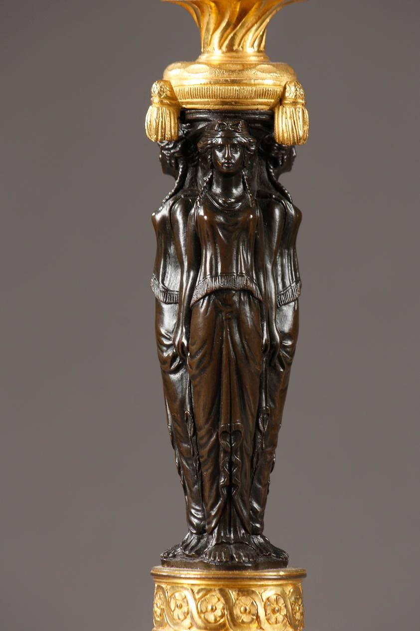 Pair of Empire bronze and marble candelabra with three branches each. The lights are supported by bronze caryatids with dark brown patina, who are dressed in long tunics à l’antique and have their backs to a pillar, forming the stem. The S-shaped